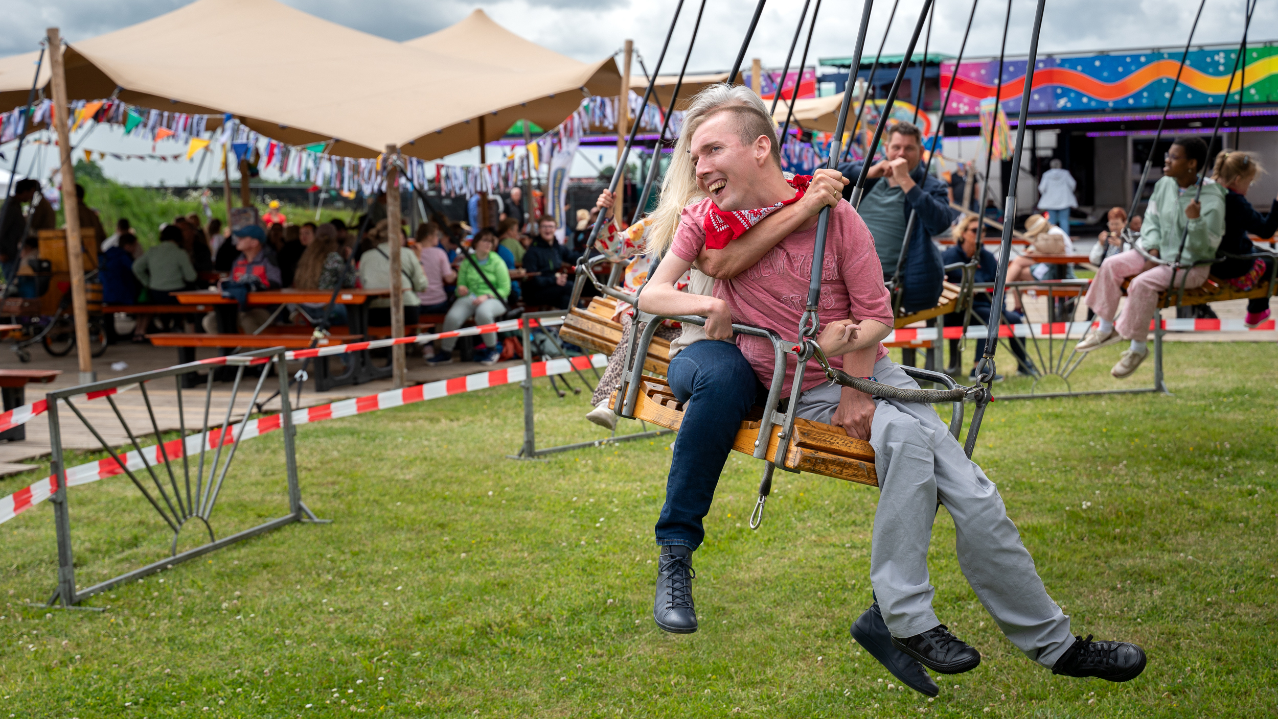 Een lachende man zit op een schommel bij een festivalterrein met kleurrijke decoraties en bezoekers op de achtergrond.