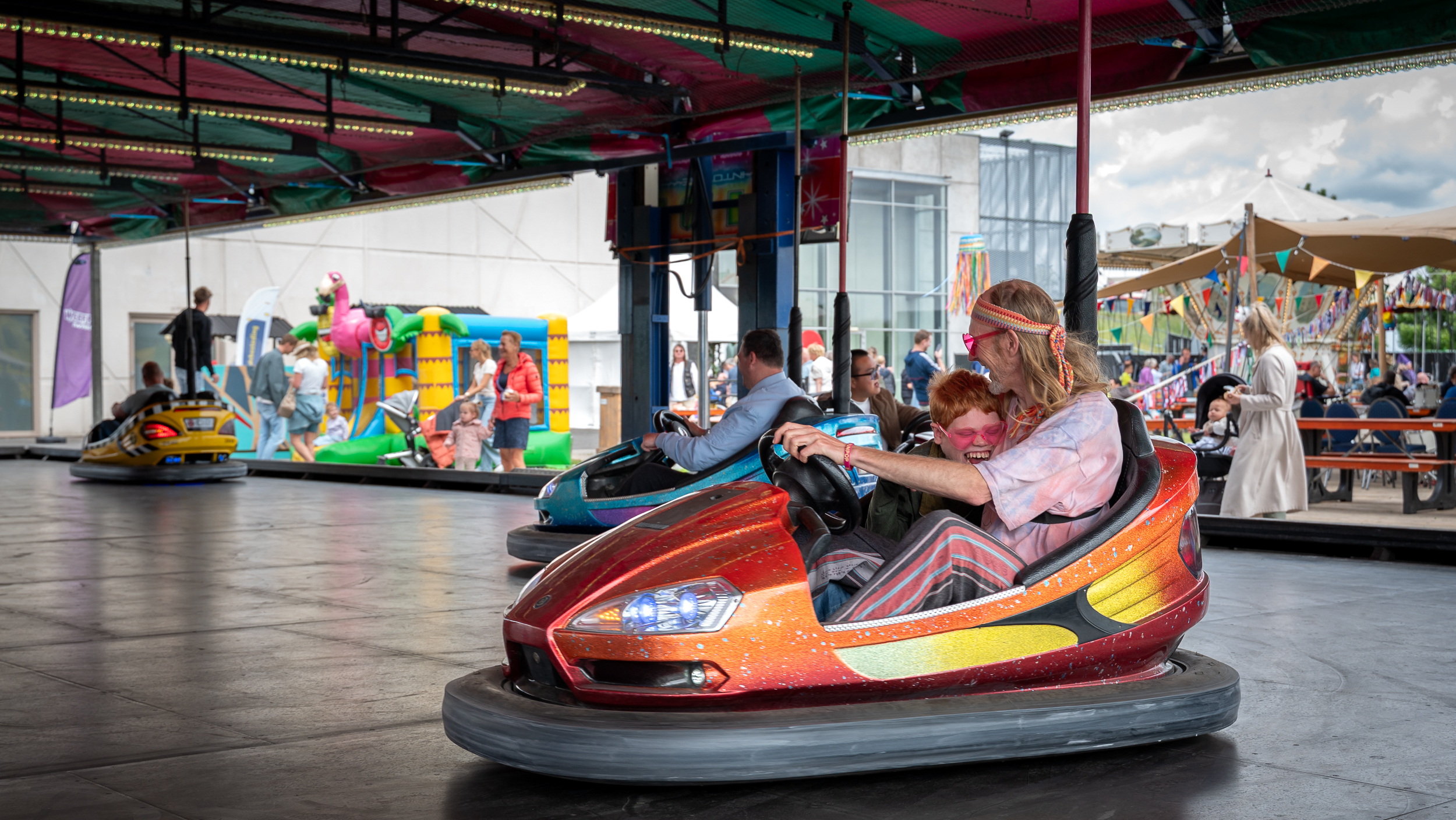 Een kind en een volwassene lachen samen in een botsauto op een kermis, omgeven door andere attracties en mensen.