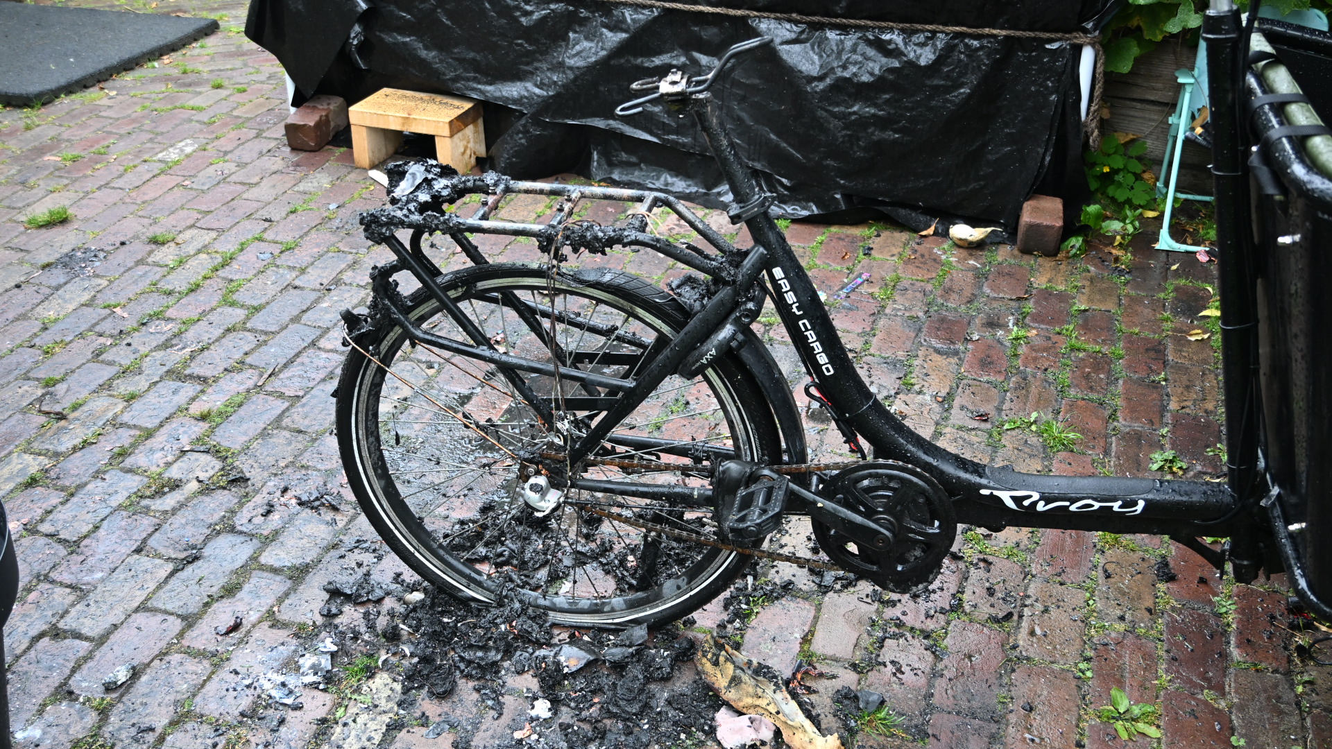 Verbrande fiets op een natte bakstenen straat, met zichtbare schade aan het frame en de wielen.