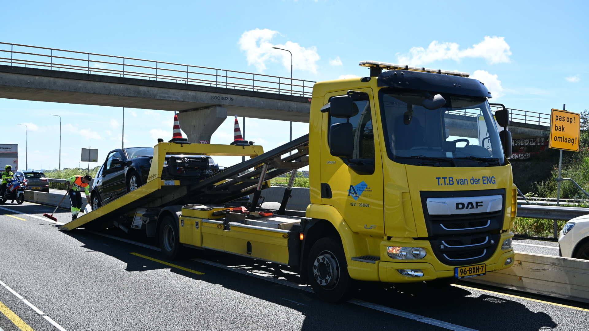 Een zwarte auto wordt door een gele sleepwagen opgeladen op een snelweg, terwijl een man in veiligheidskleding de weg veegt.