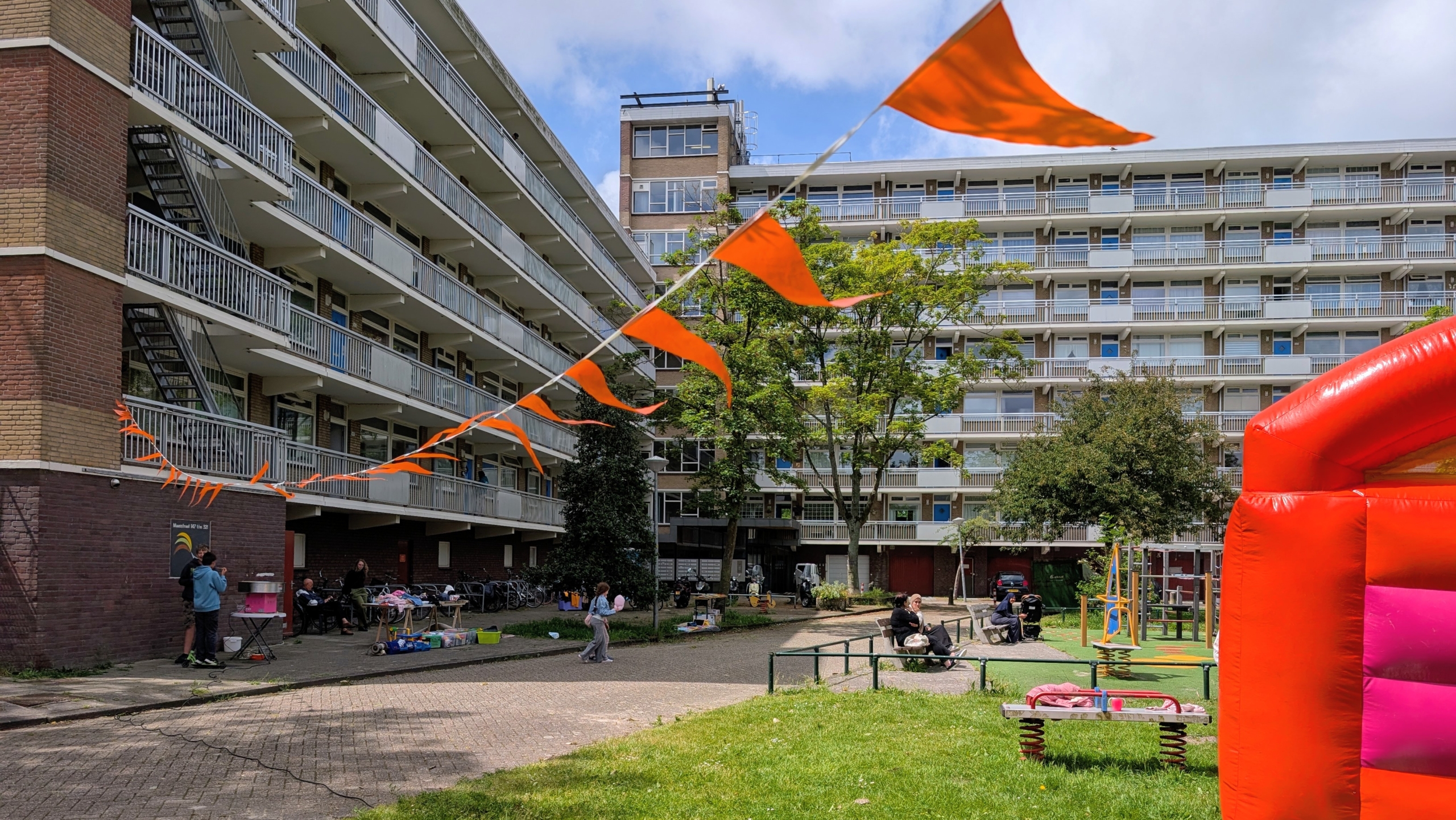 Flatgebouw met balkons en oranje vlaggetjes, mensen op een plein en een felgekleurde speelstructuur op de voorgrond.