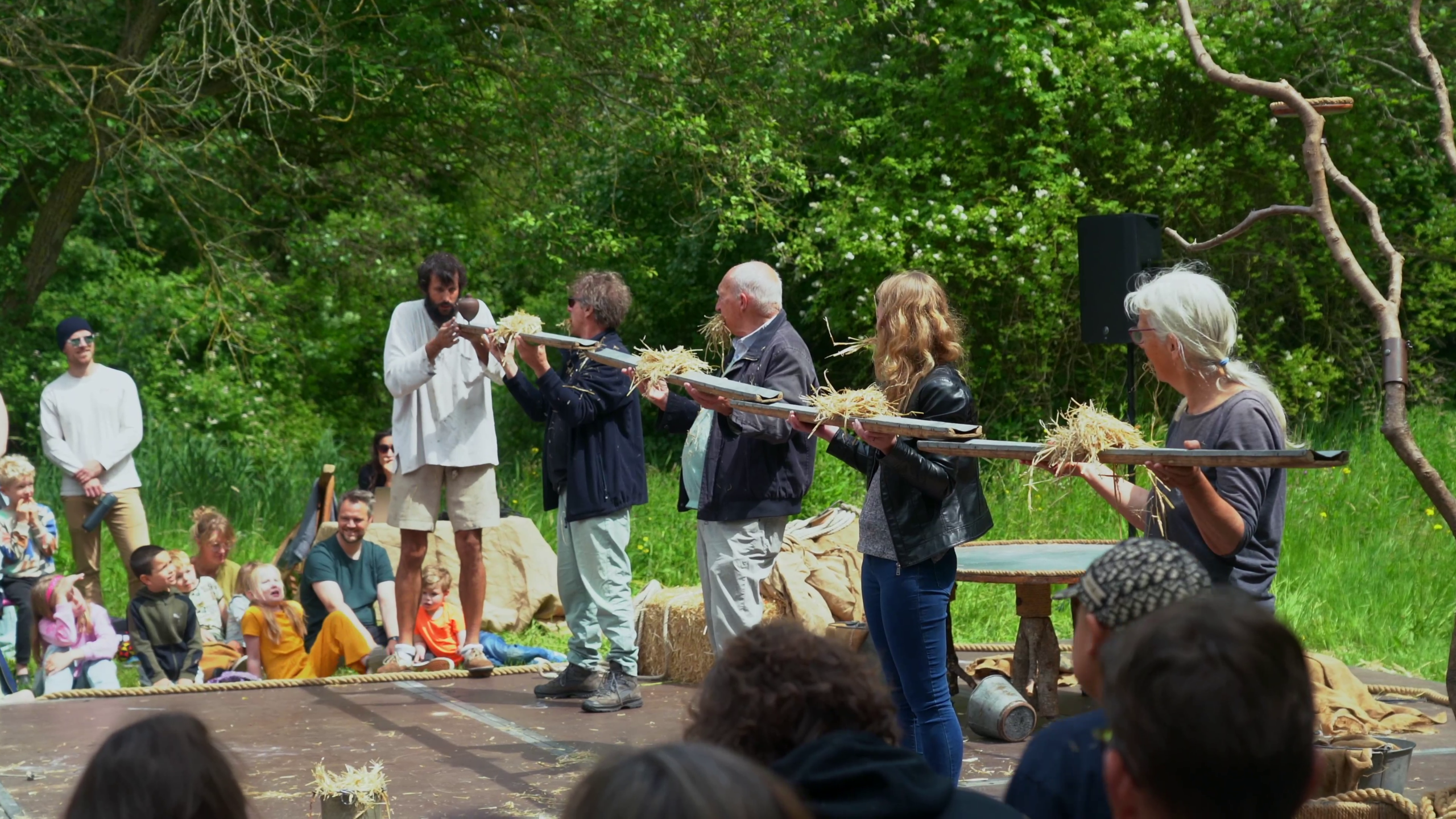 Groep mensen in openluchtvoorstelling met hooi op lange planken; toeschouwers kijken geboeid toe.