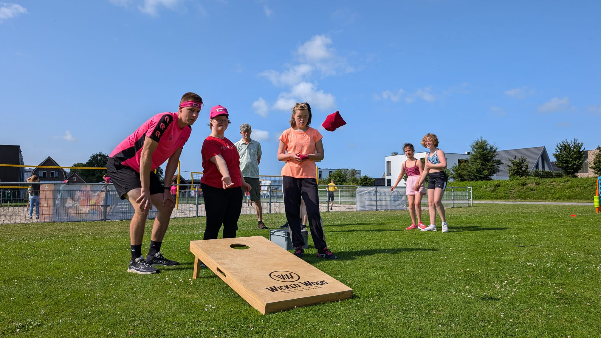 Mensen spelen cornhole op een grasveld; een rood zakje wordt naar het bord gegooid onder een blauwe hemel.