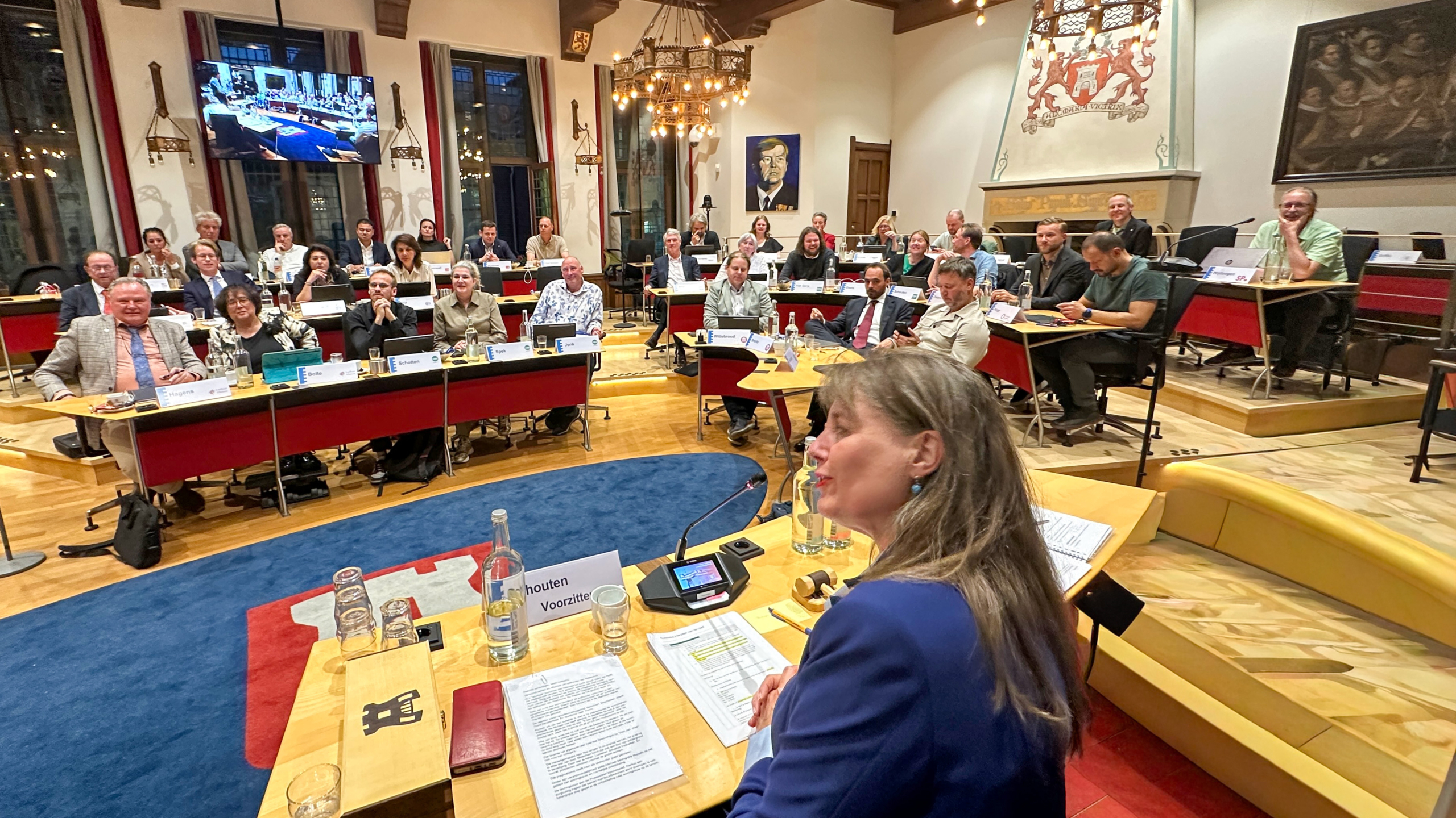 Een bestuursvergadering met deelnemers zittend aan tafels in een grote kamer met een kroonluchter en kunst aan de muren.