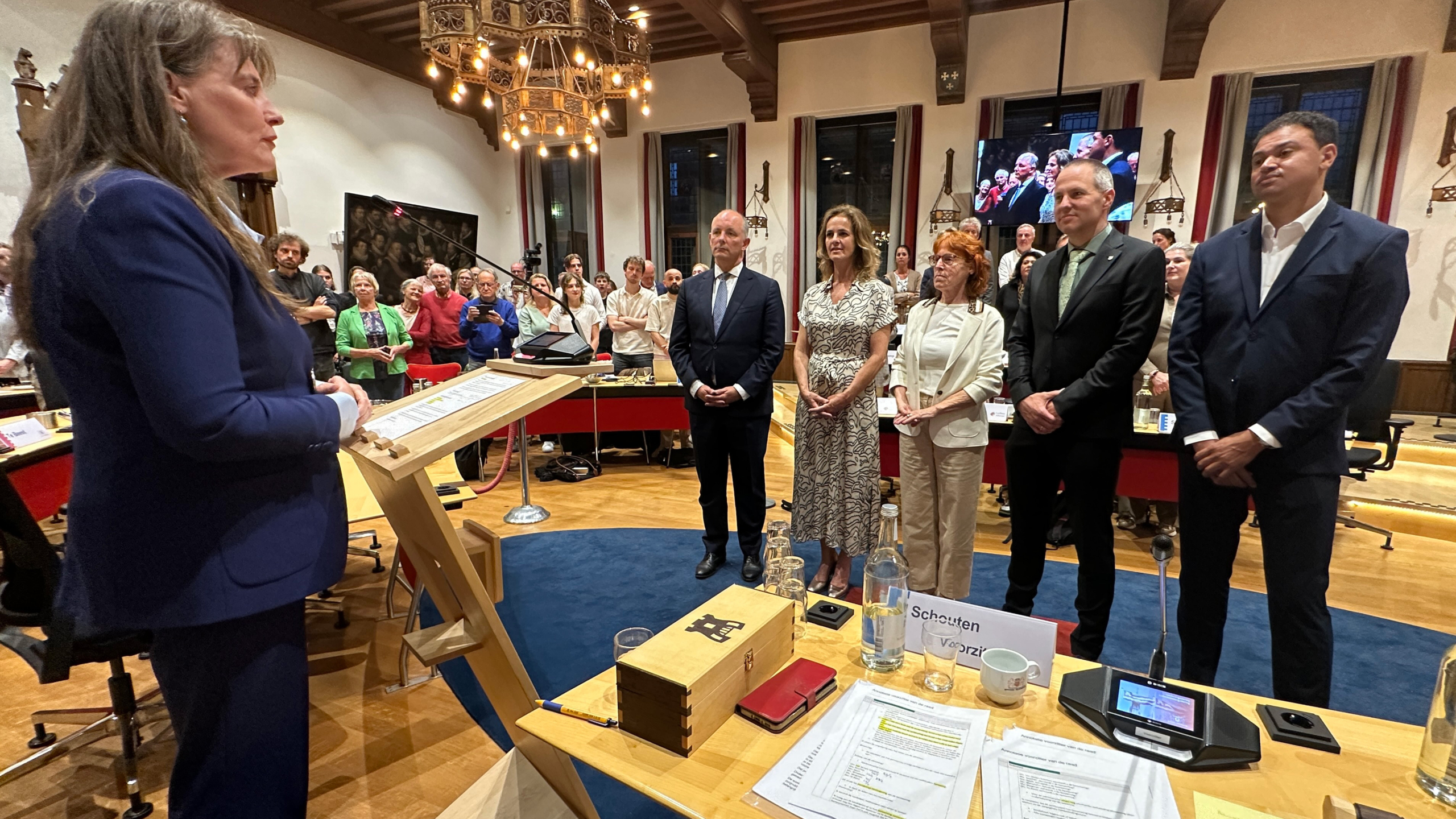 Een vrouw in een zaal staat achter een lessenaar en spreekt tot een groep mensen, die aandachtig luisteren.