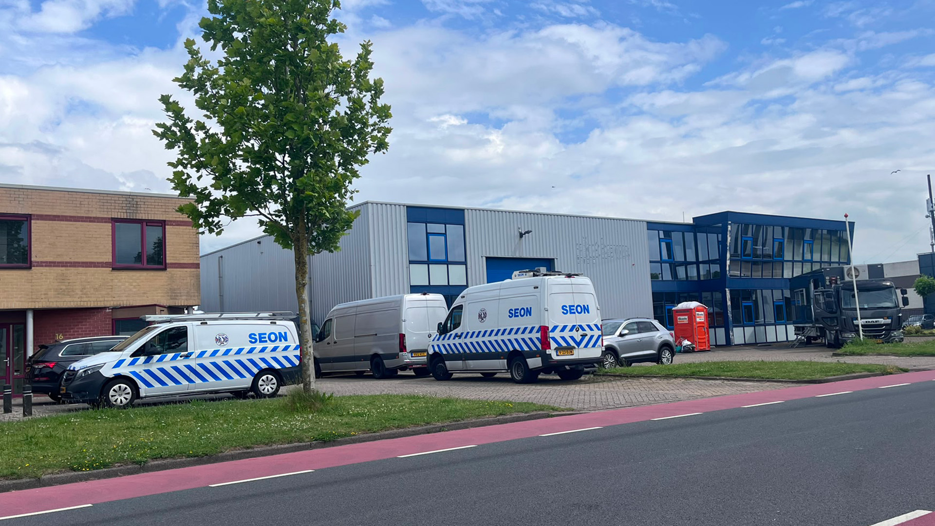 Witte bestelwagens van SEON geparkeerd voor een grijs industrieel gebouw met een boom op de voorgrond.