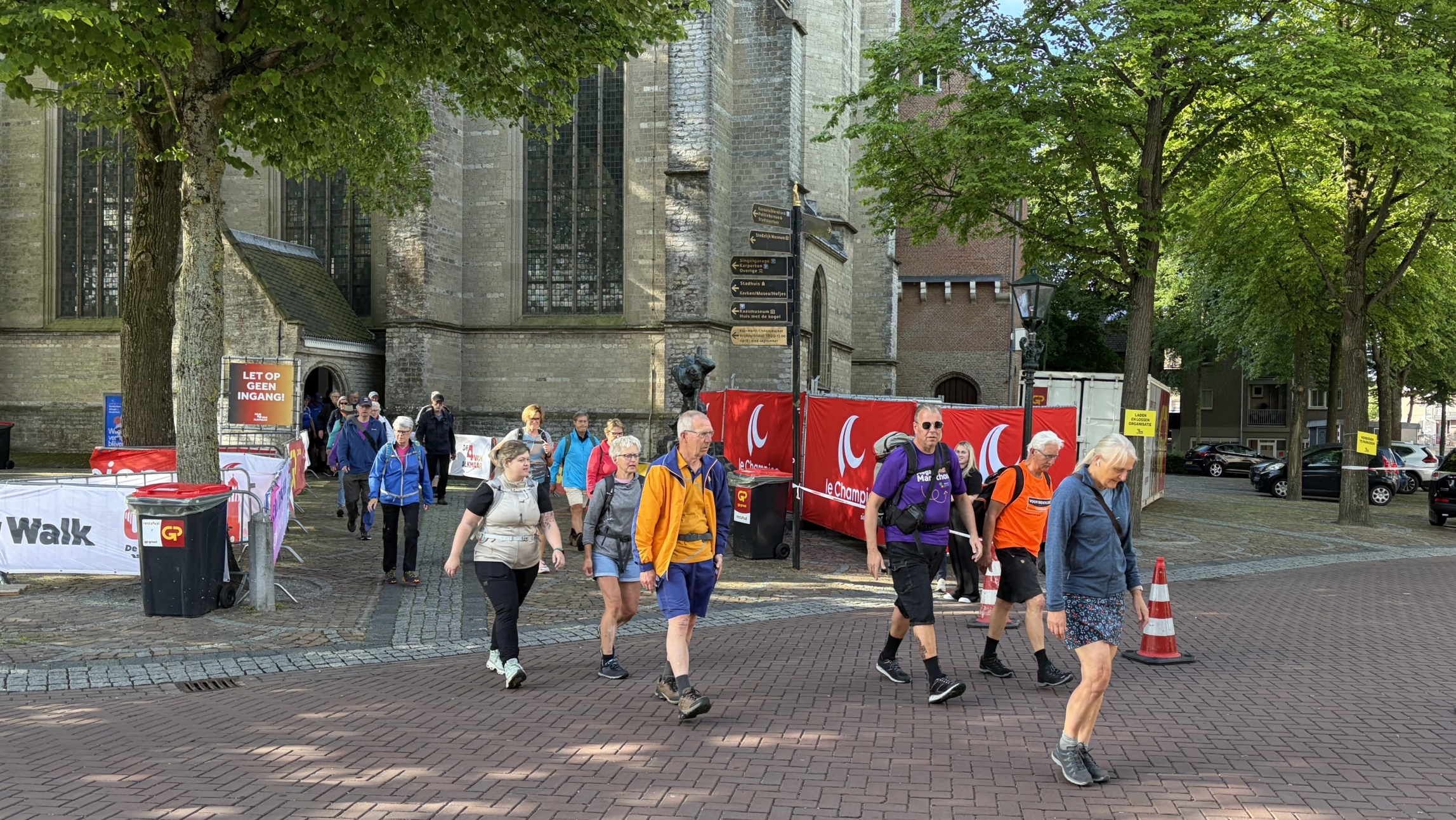 Mensen lopen over een geplaveide straat naast een historische kerk met bomen en verkeerskegels in de buurt.