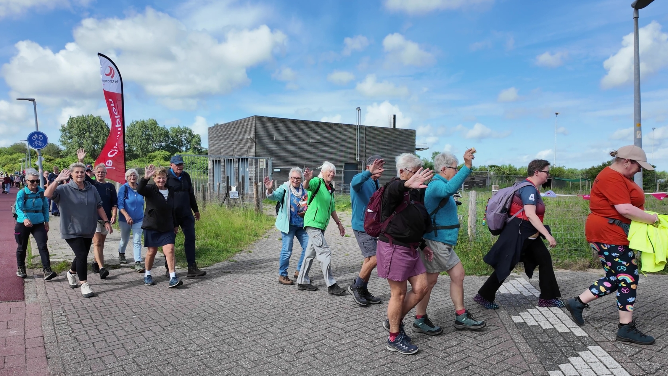 Een groep mensen wandelt over een stoep langs een fietspad, sommige zwaaien naar de camera op een zonnige dag.