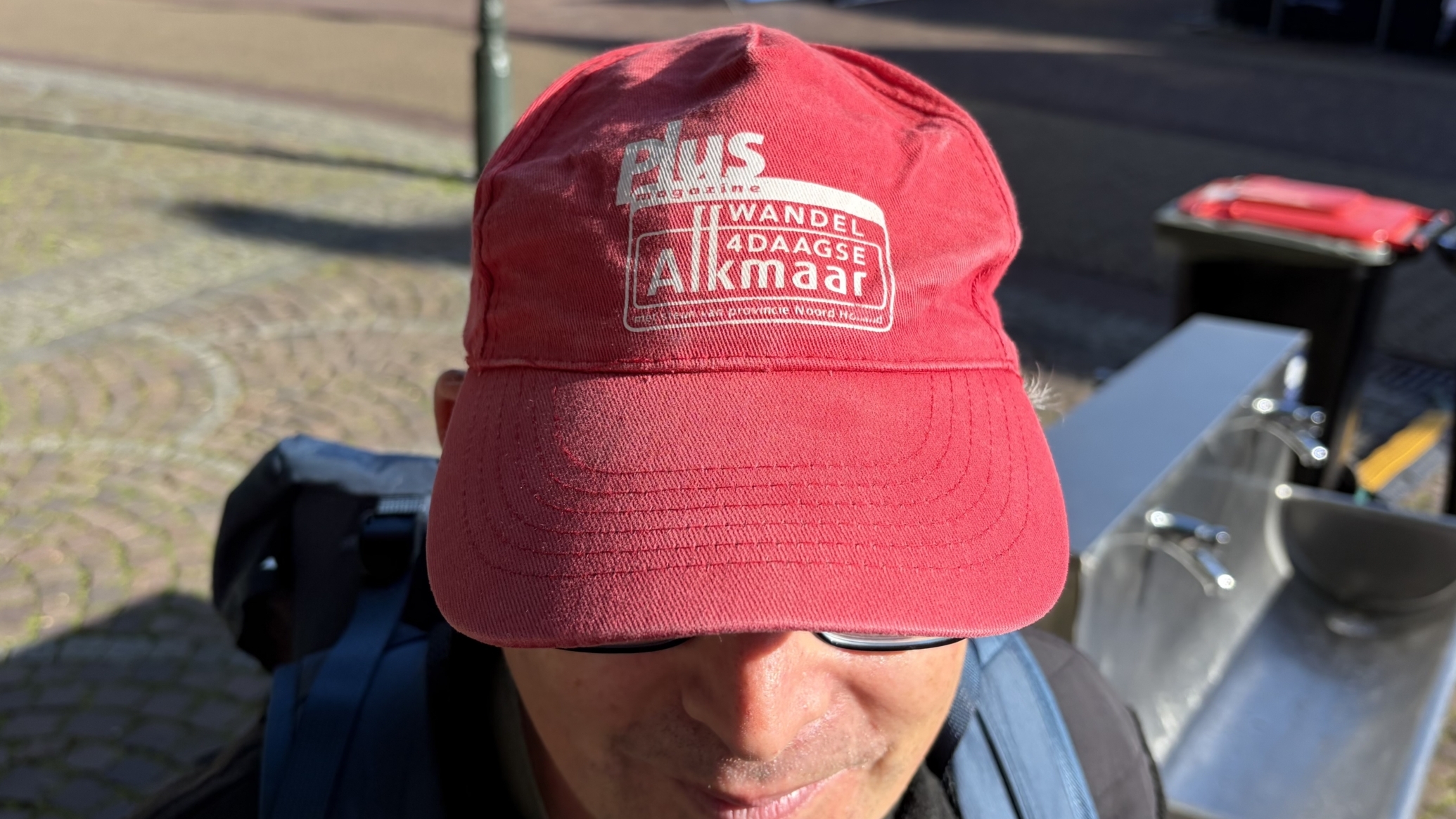 Man met een rode pet met de tekst "Wandelvierdaagse Alkmaar" staat buiten met een rugzak.