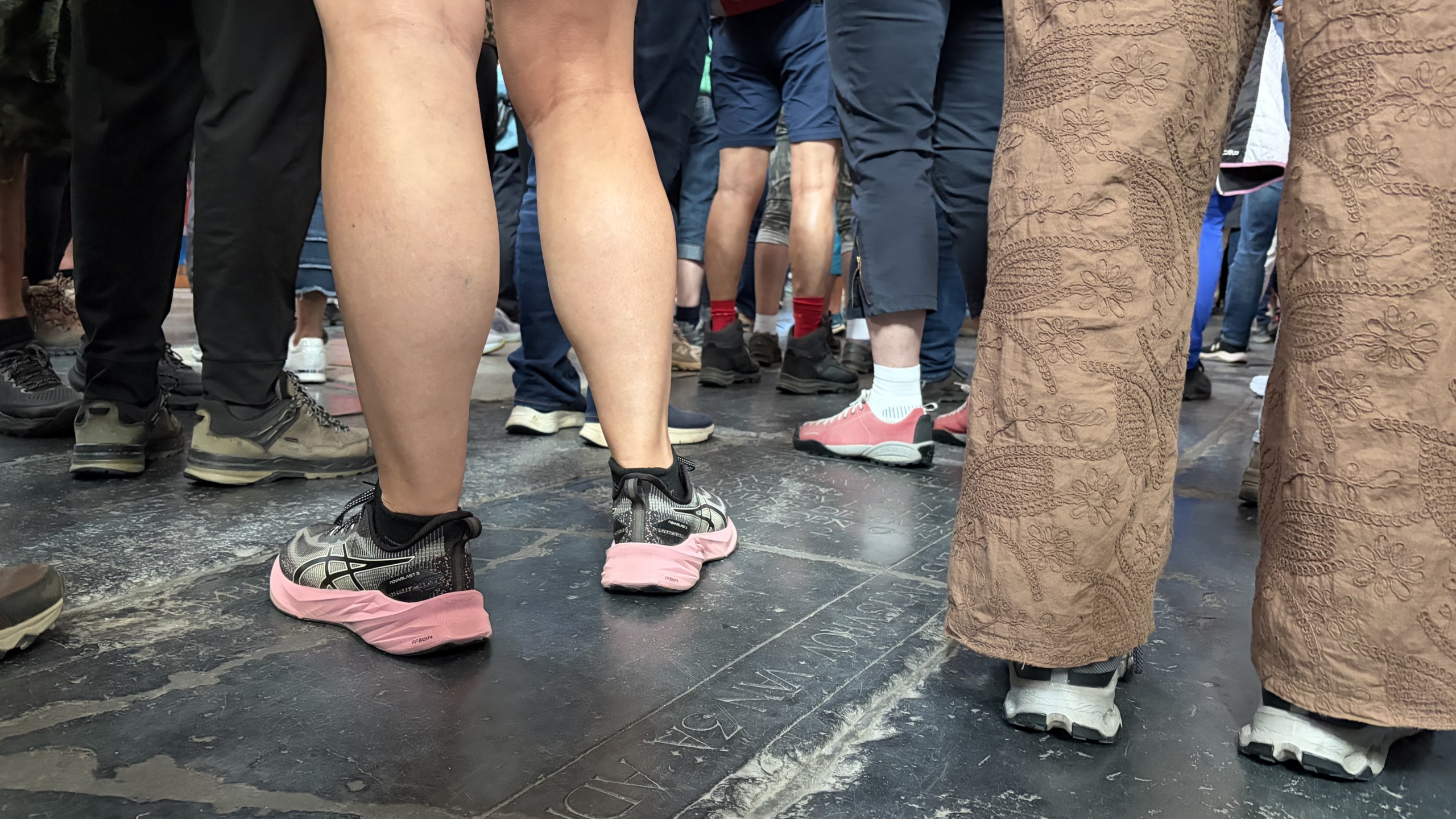 Een groep mensen in sportschoenen en casual kledingstijlen staat dicht bij elkaar op een stenen vloer met inscripties.