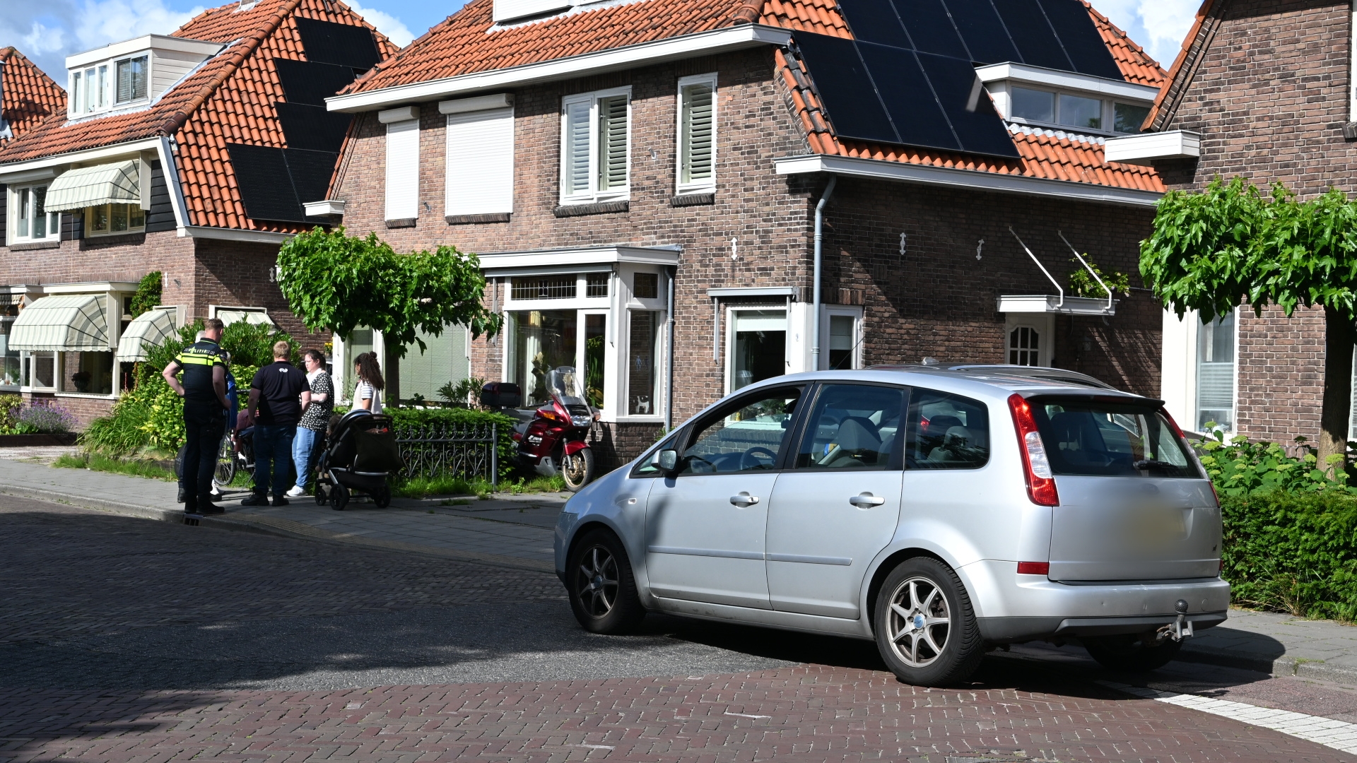 Politieagent en meerdere mensen staan naast een zilveren auto in een woonwijk met bakstenen huizen en rode daken.