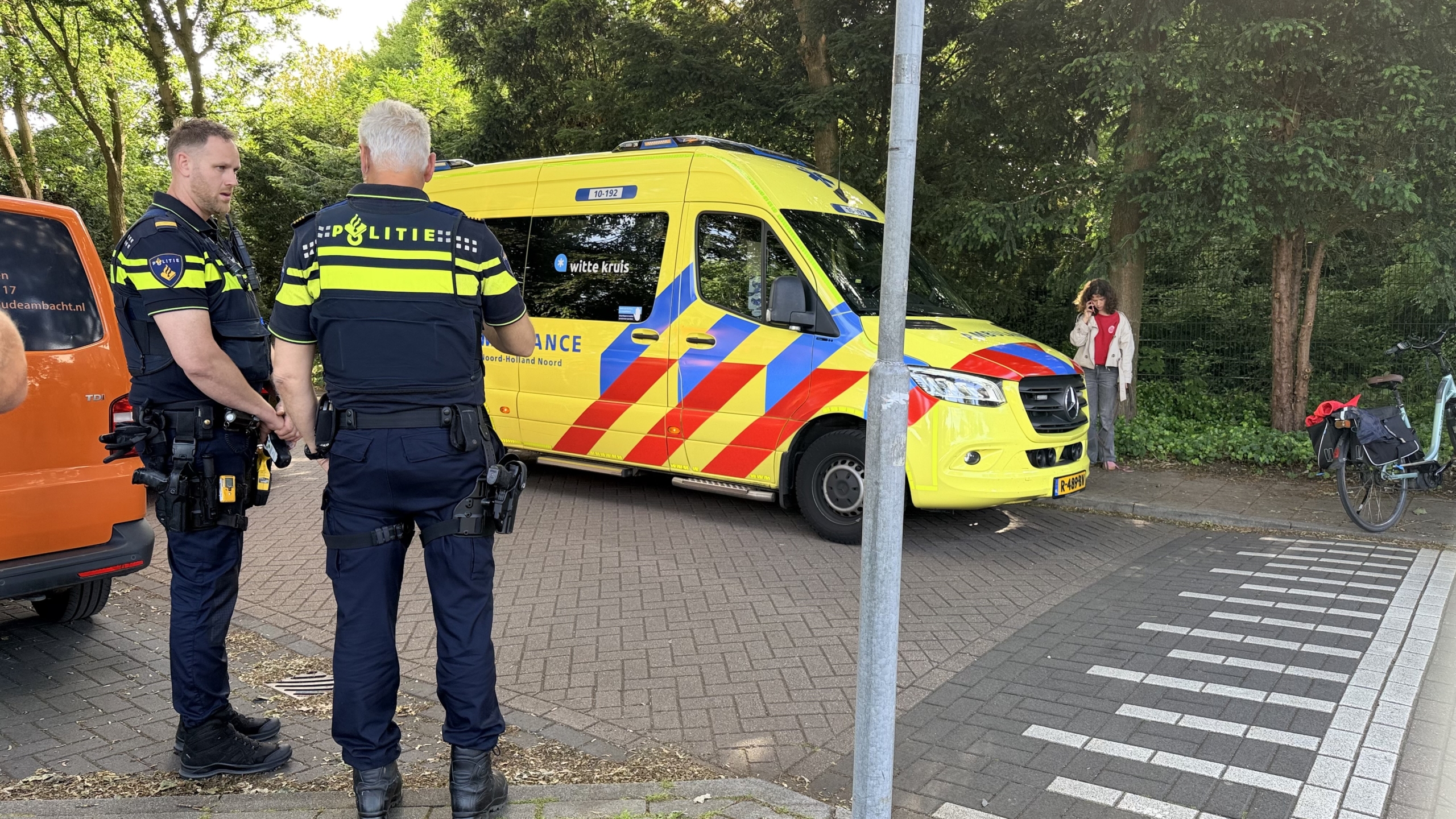 Twee politieagenten staan bij een geparkeerde ambulance, terwijl een vrouw verderop aan de telefoon is.