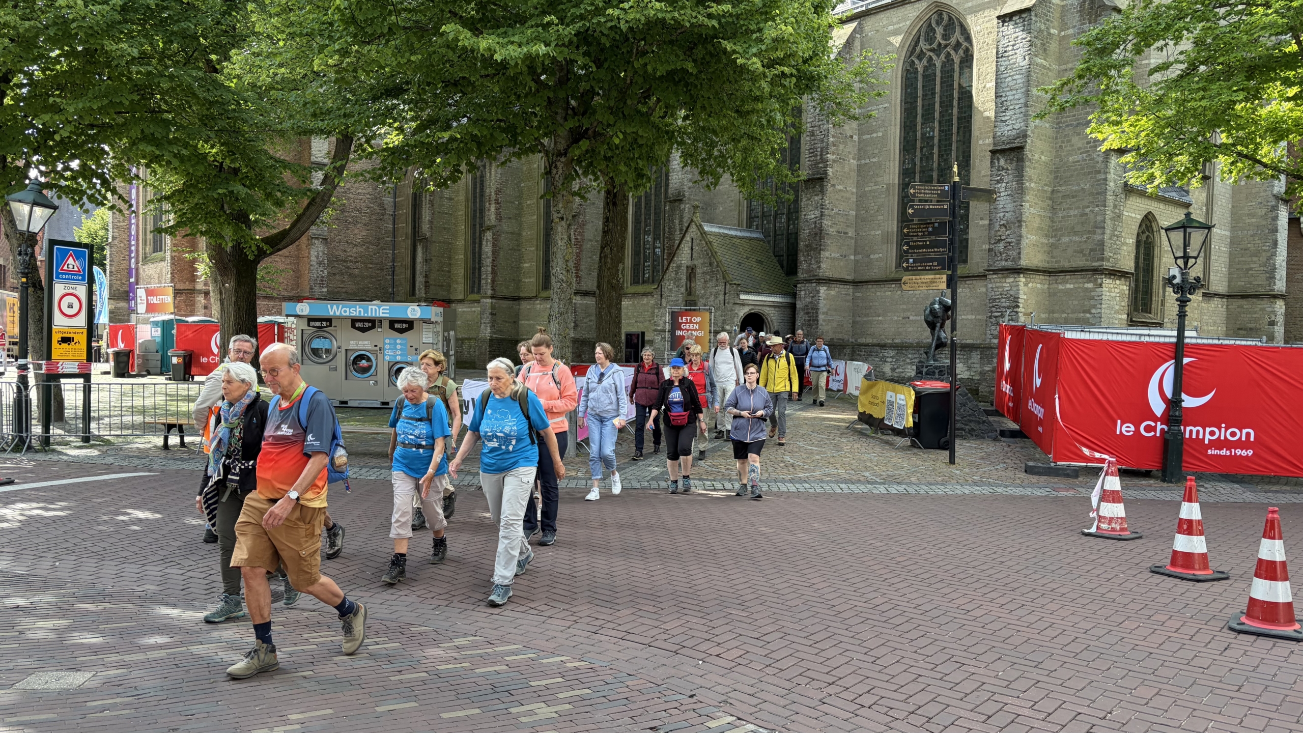 Groep wandelaars loopt over een plein langs een grote kerk met rode hekken en borden op de achtergrond.