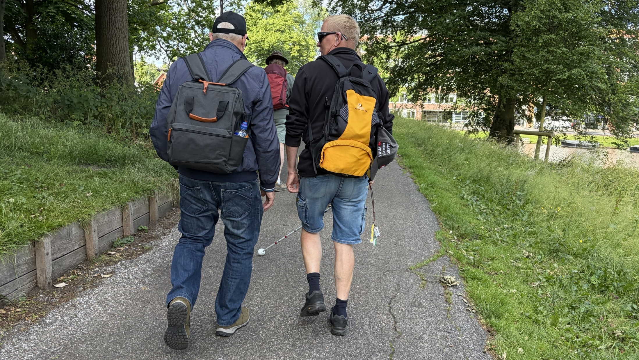Twee mannen met rugzakken lopen over een pad langs een grasveld en een waterkanaal, omringd door bomen.