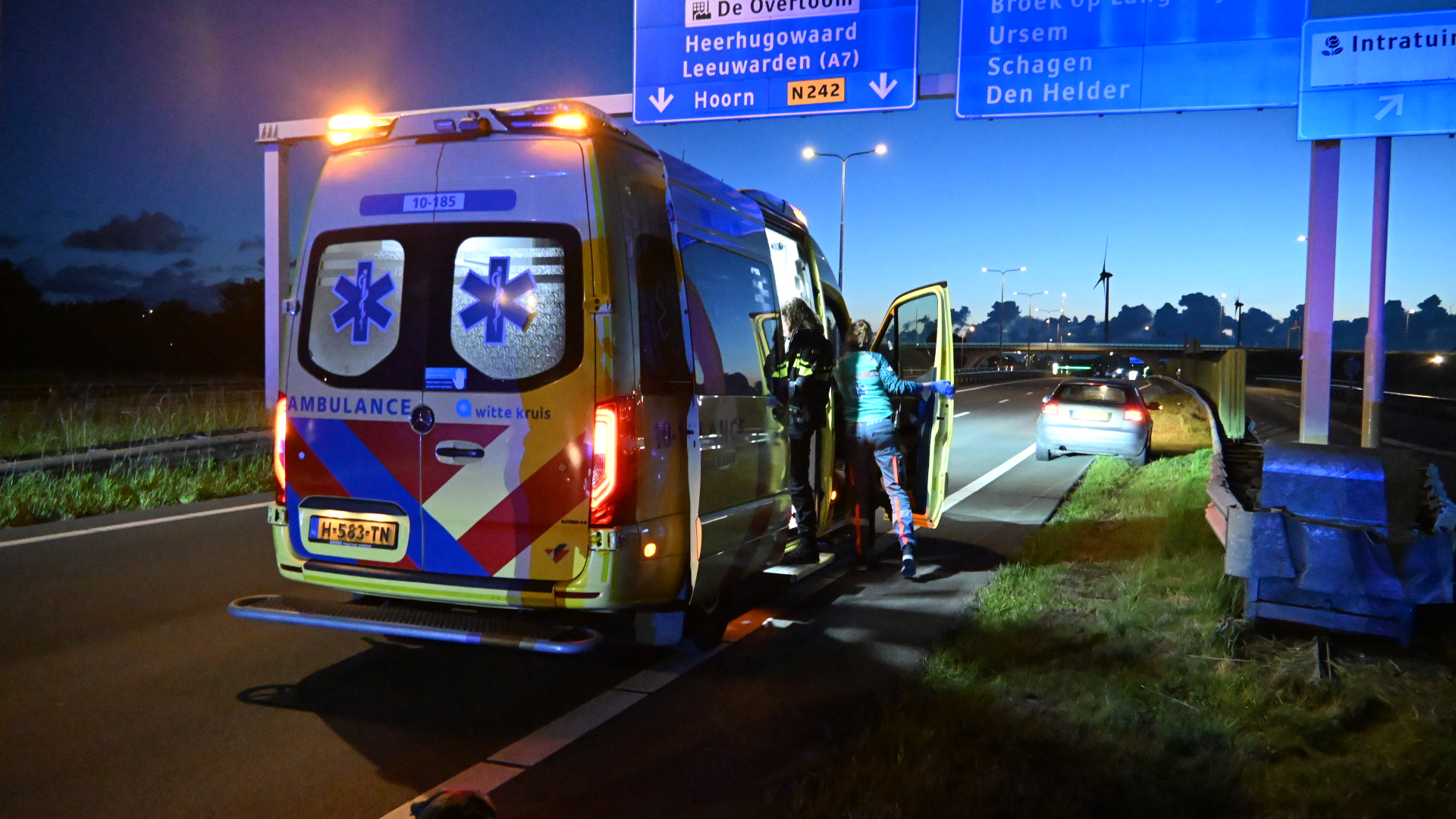 Ambulance met geopende deuren langs de weg in de schemering, naast een auto en verkeersbord.