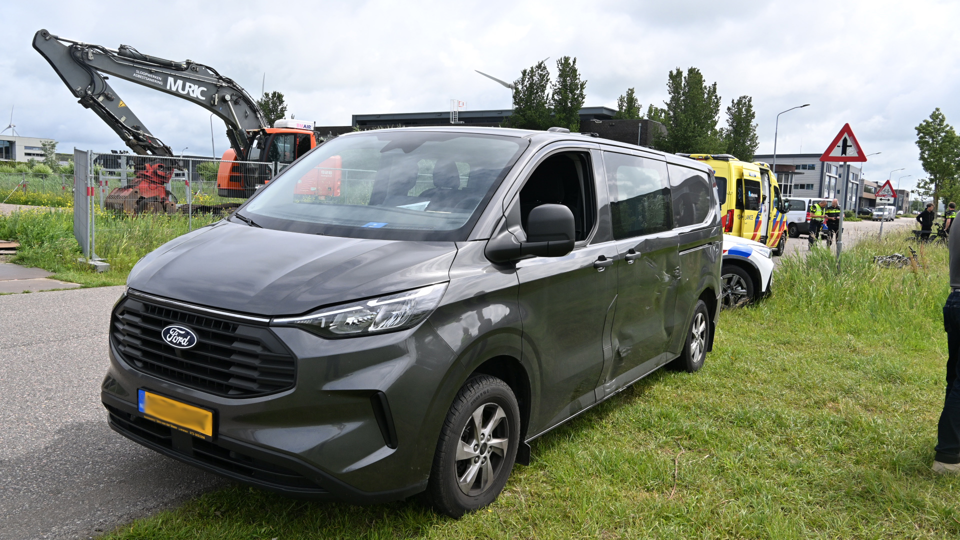 Een grijze bestelwagen staat geparkeerd naast een bouwterrein met een graafmachine, naast een ambulance en agenten.