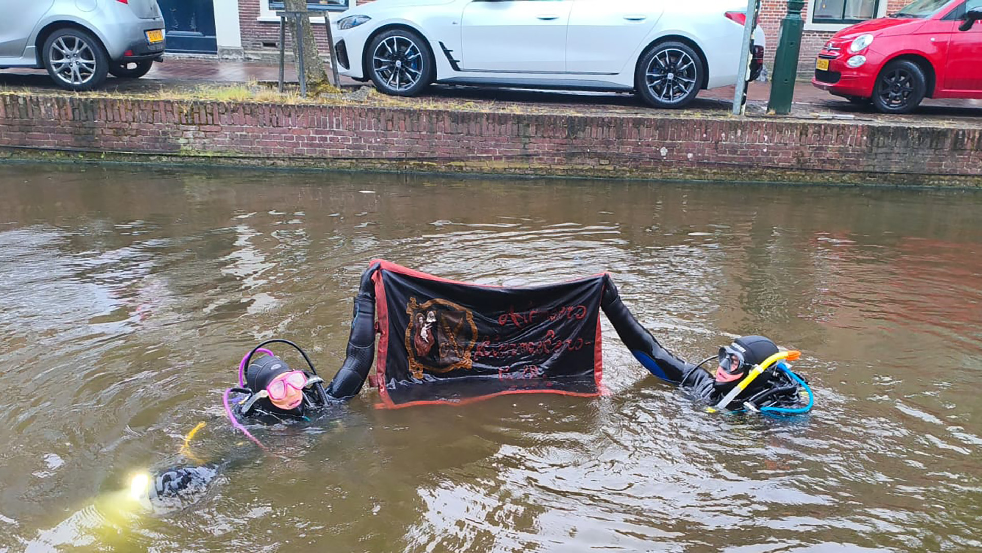 Twee duikers in een kanaal houden een spandoek met een ontwerp en tekst omhoog. Op de achtergrond geparkeerde auto's.