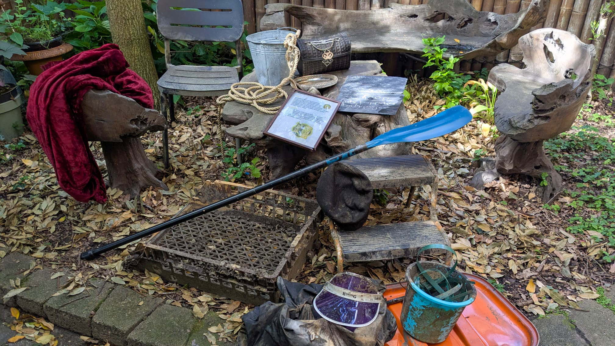 Een tuinsetting met houtgesneden meubels, een blauwe peddel, een touw, een metalen emmer en bladeren op de grond.