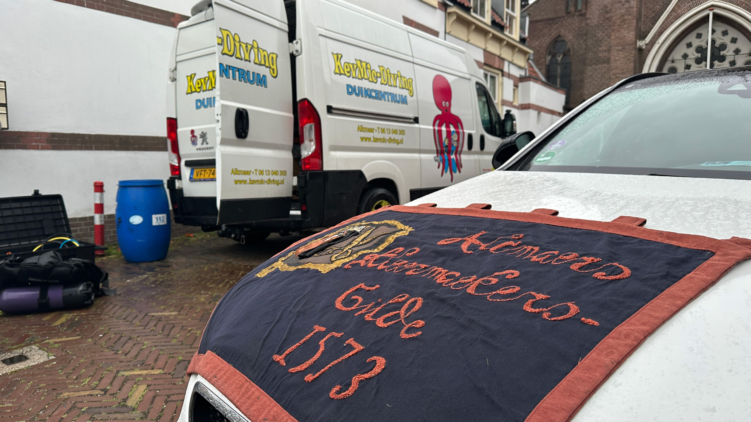 Bestelbus met duikcentrumlogo naast een auto met een doek waarop 'Alkmaerder Gilde 1573' staat, geparkeerd bij een gebouw.