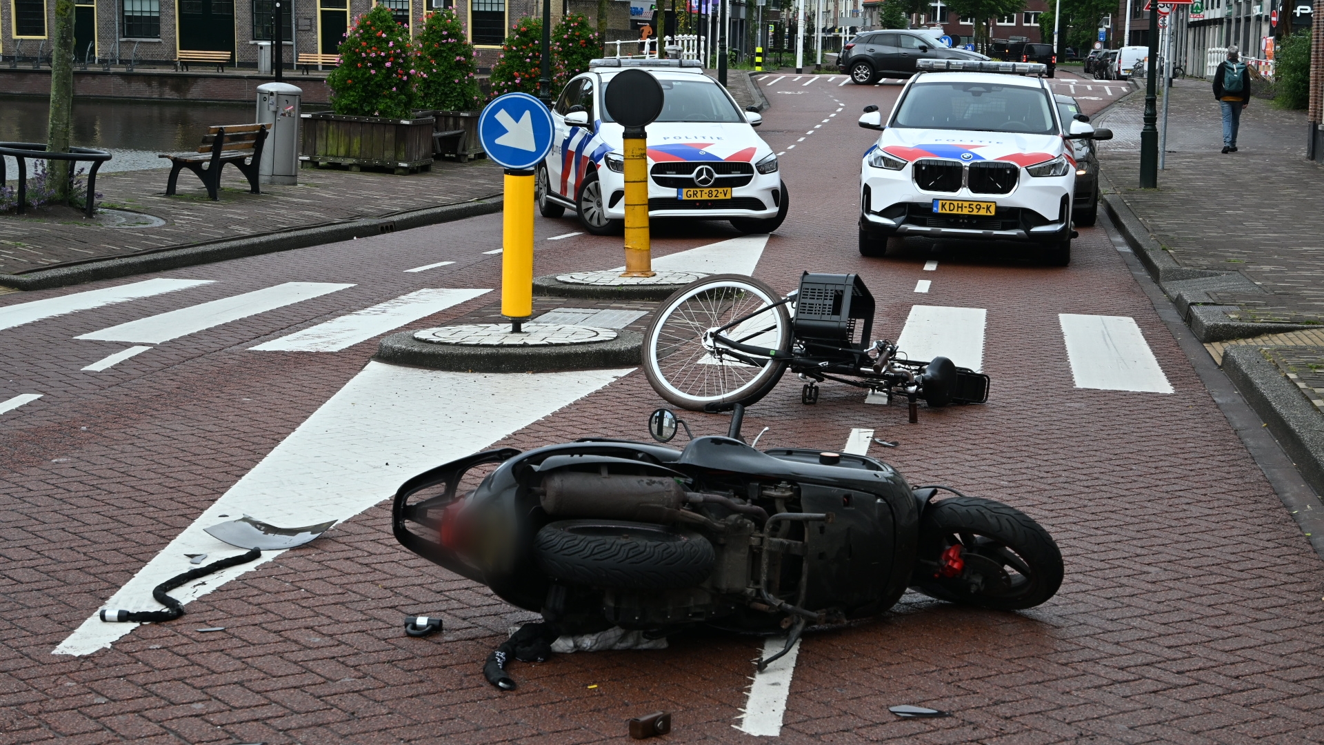Omgevallen scooter en fiets op de weg naast politieauto's in een stedelijke omgeving.