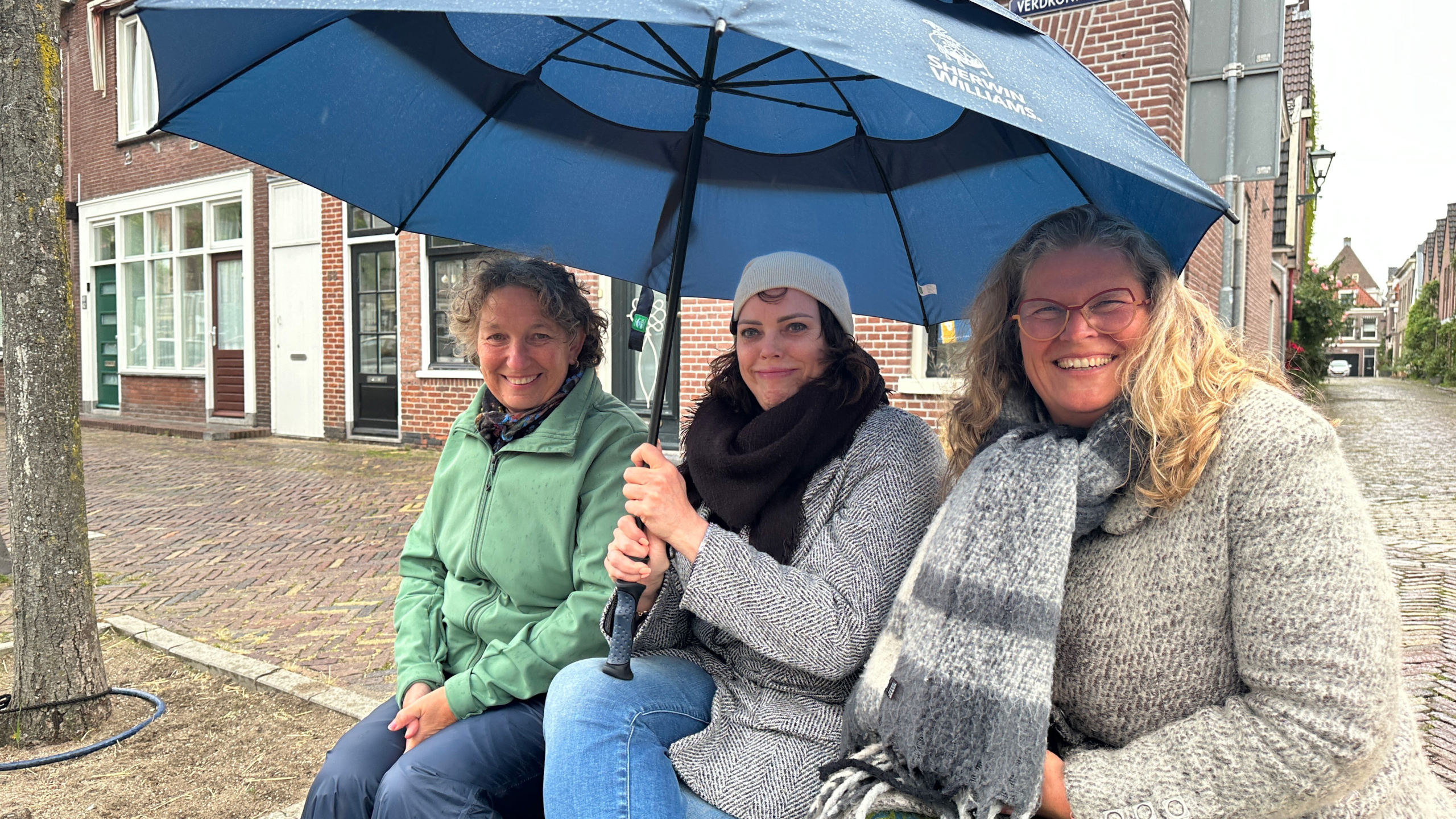 Drie vrouwen zitten op een bank met een blauwe paraplu, terwijl het buiten regent. Ze dragen warme jassen en sjaals.