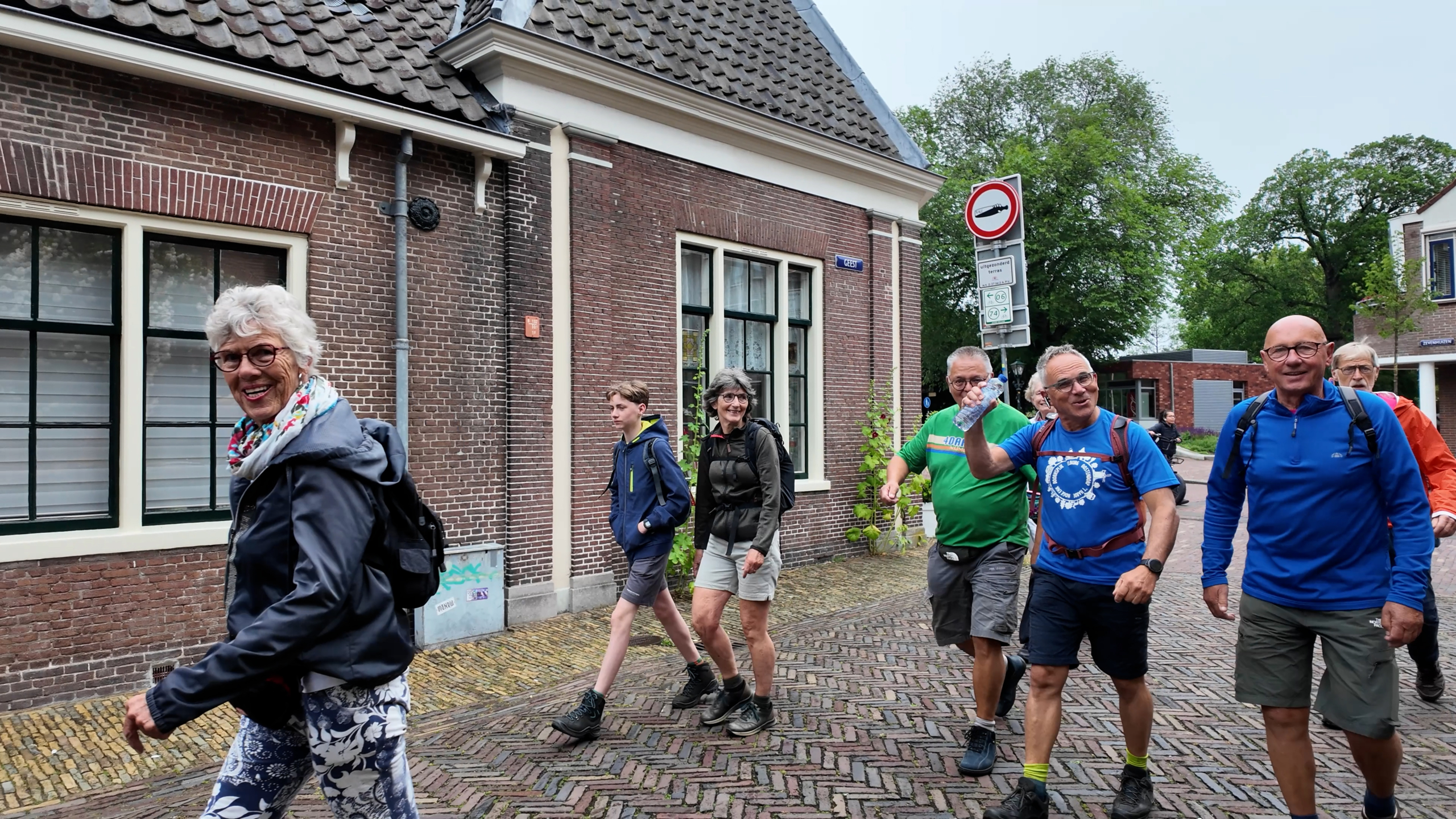 Groep wandelaars loopt vrolijk door een straat met bakstenen huizen en groen omlijst vensters, op een bewolkte dag.