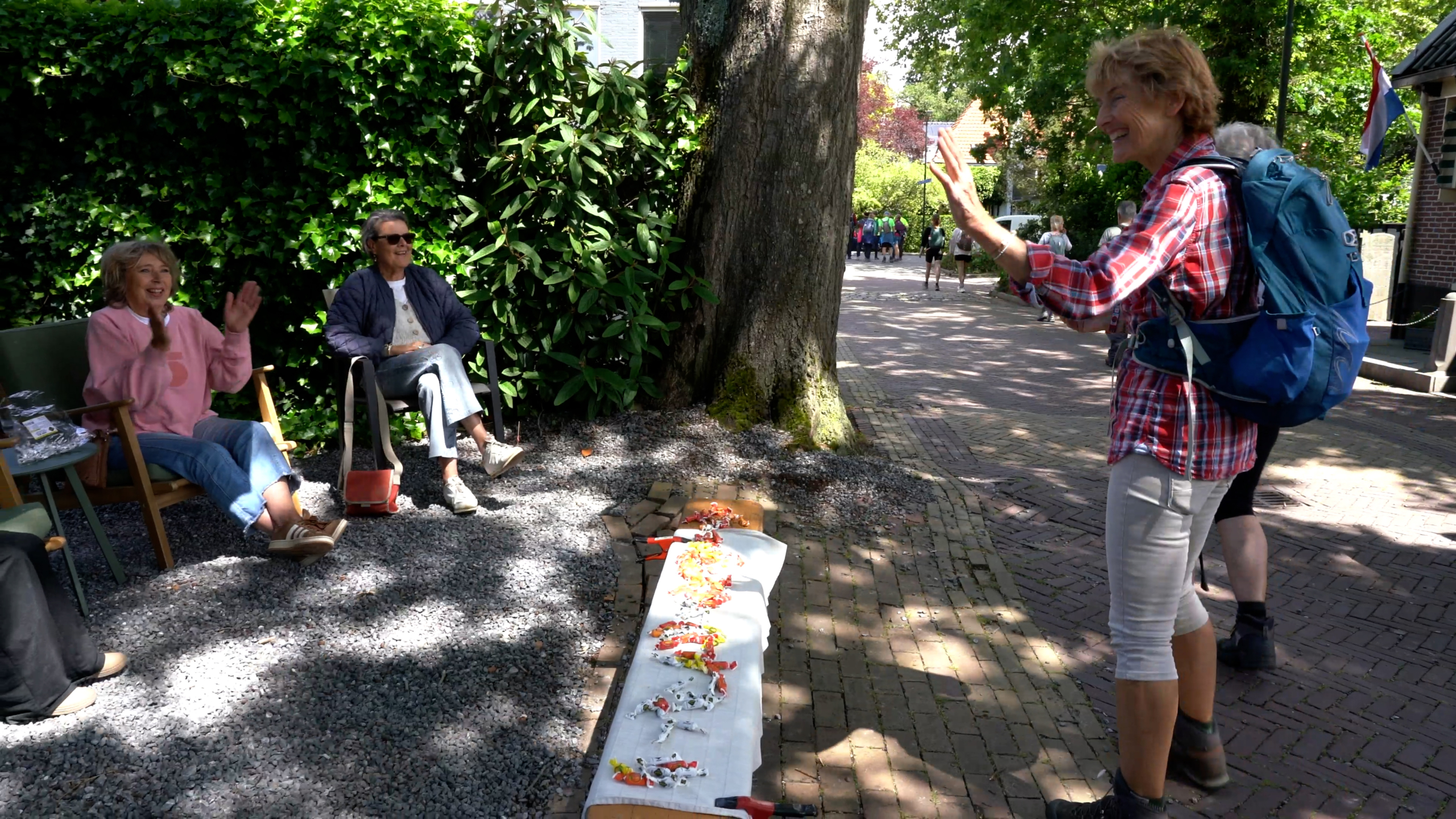 Een vrouw met een rugzak begroet twee zittende mensen in een tuin. Ze lachen en klappen.