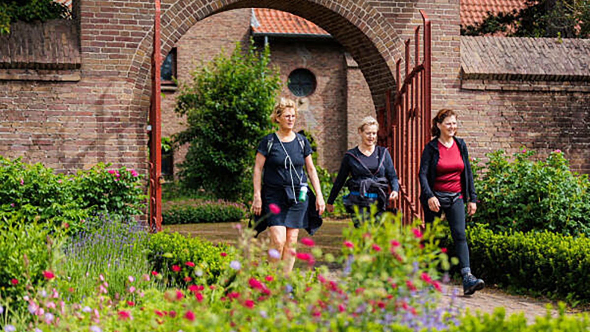 Drie vrouwen wandelen door een poort in een tuin met kleurrijke bloemen en groene struiken.