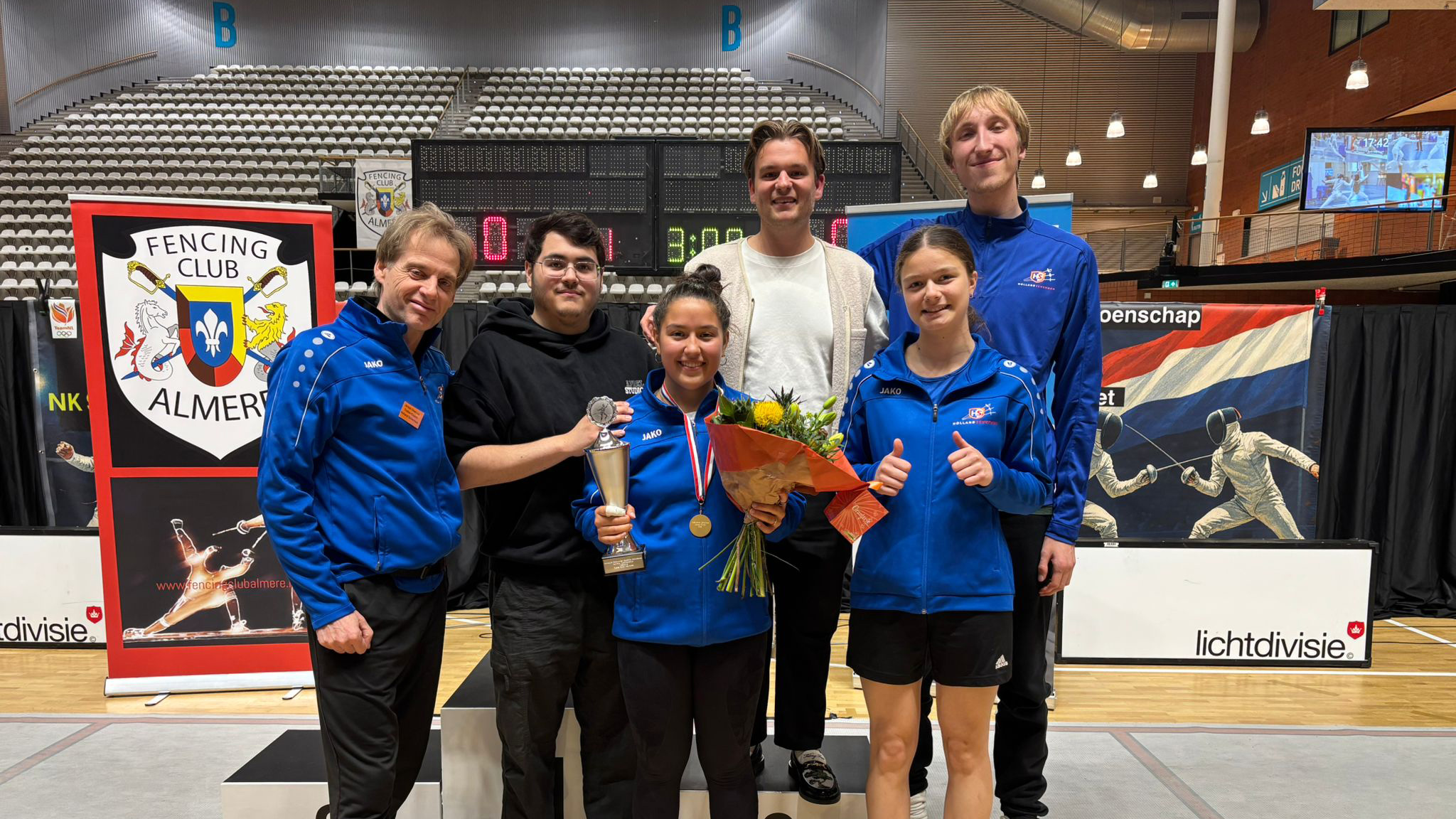 Een groep mensen poseert in een sporthal, met een vrouw in het midden die een trofee en bloemen vasthoudt. Ze dragen sportkleding.