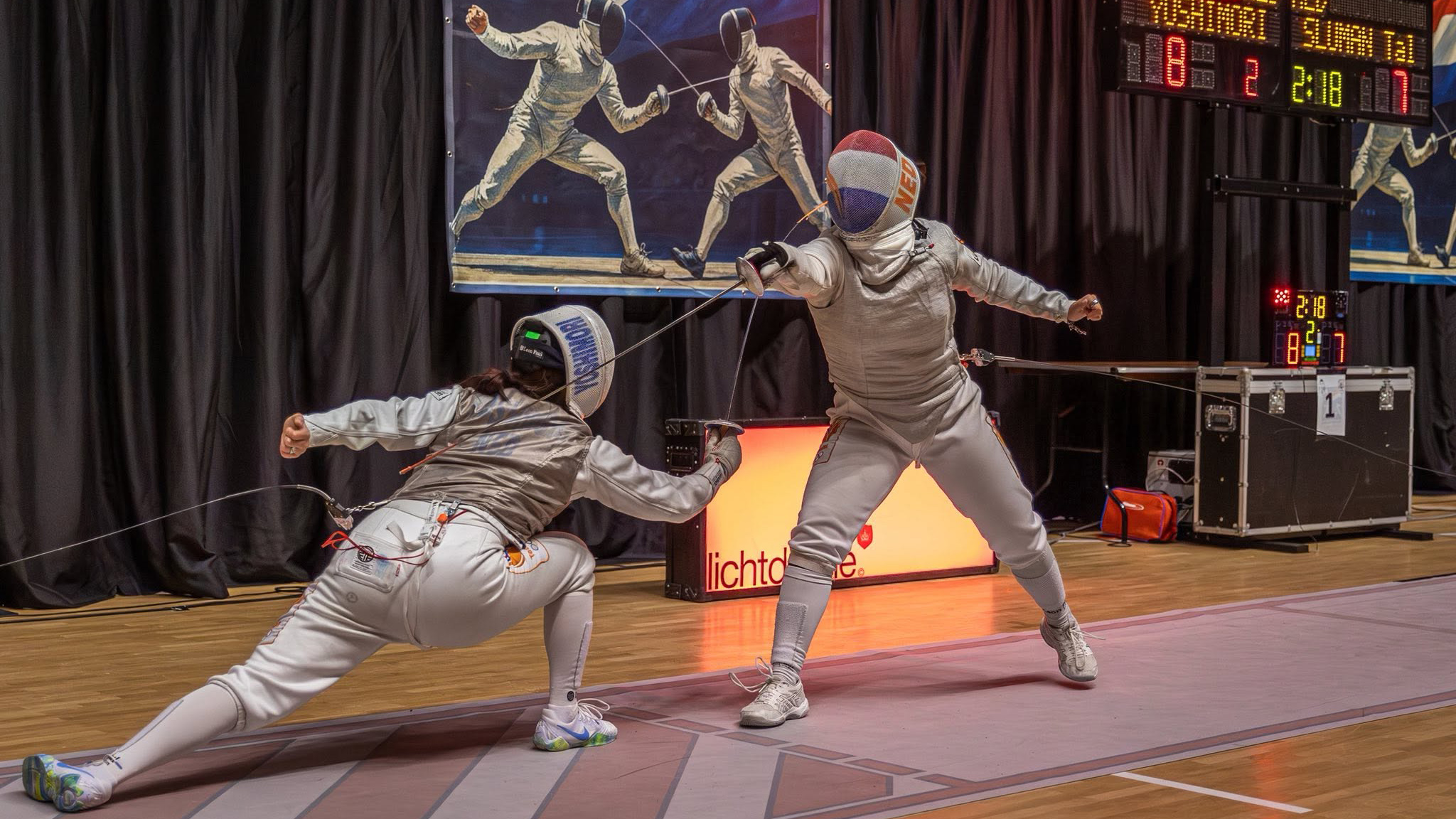 Twee schermers in actie op een indoor wedstrijd, gekleed in witte schermuitrusting met maskers.