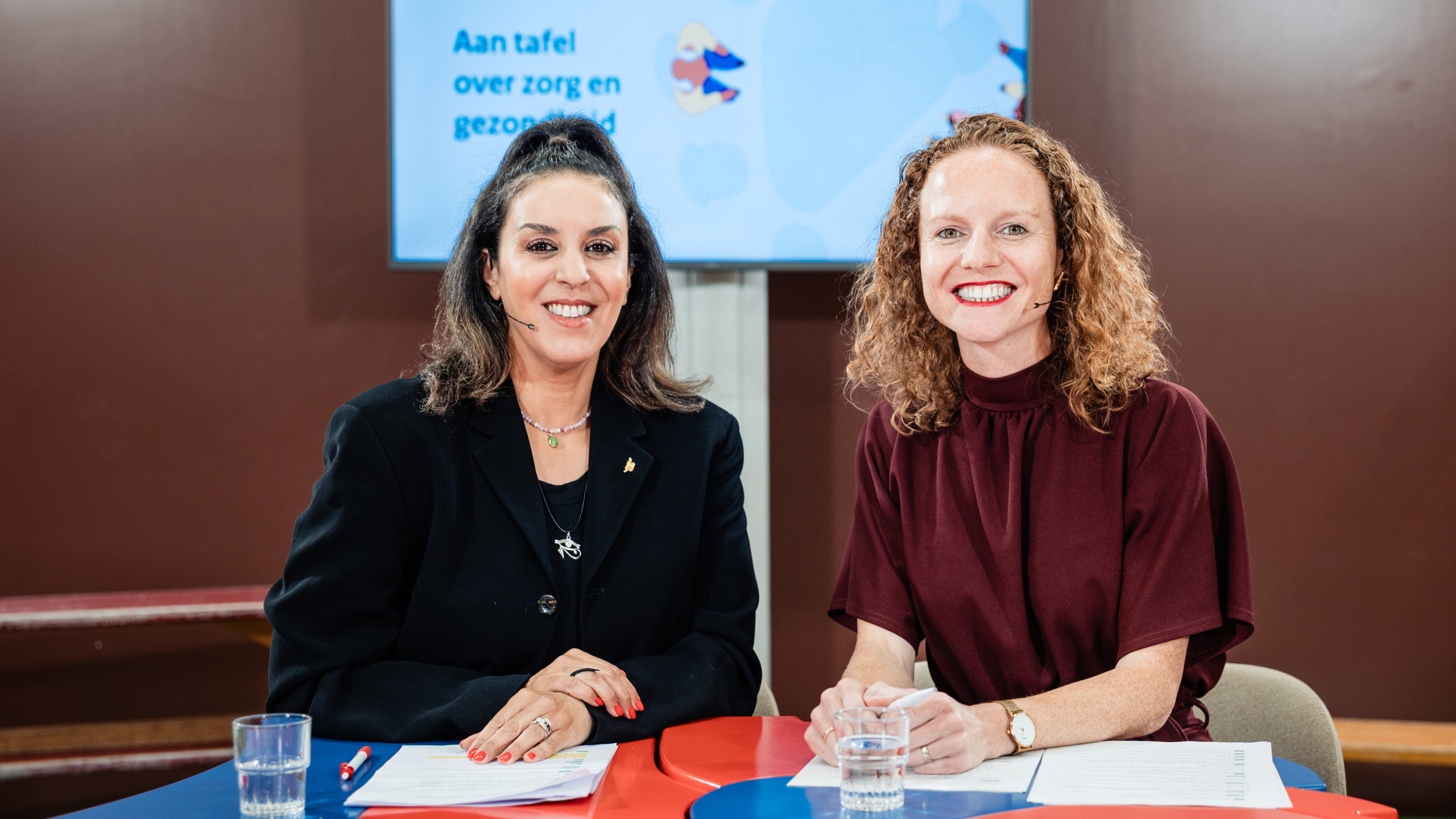 Twee vrouwen zitten aan een tafel en glimlachen naar de camera, met op de achtergrond een scherm met tekst over zorg en gezondheid.