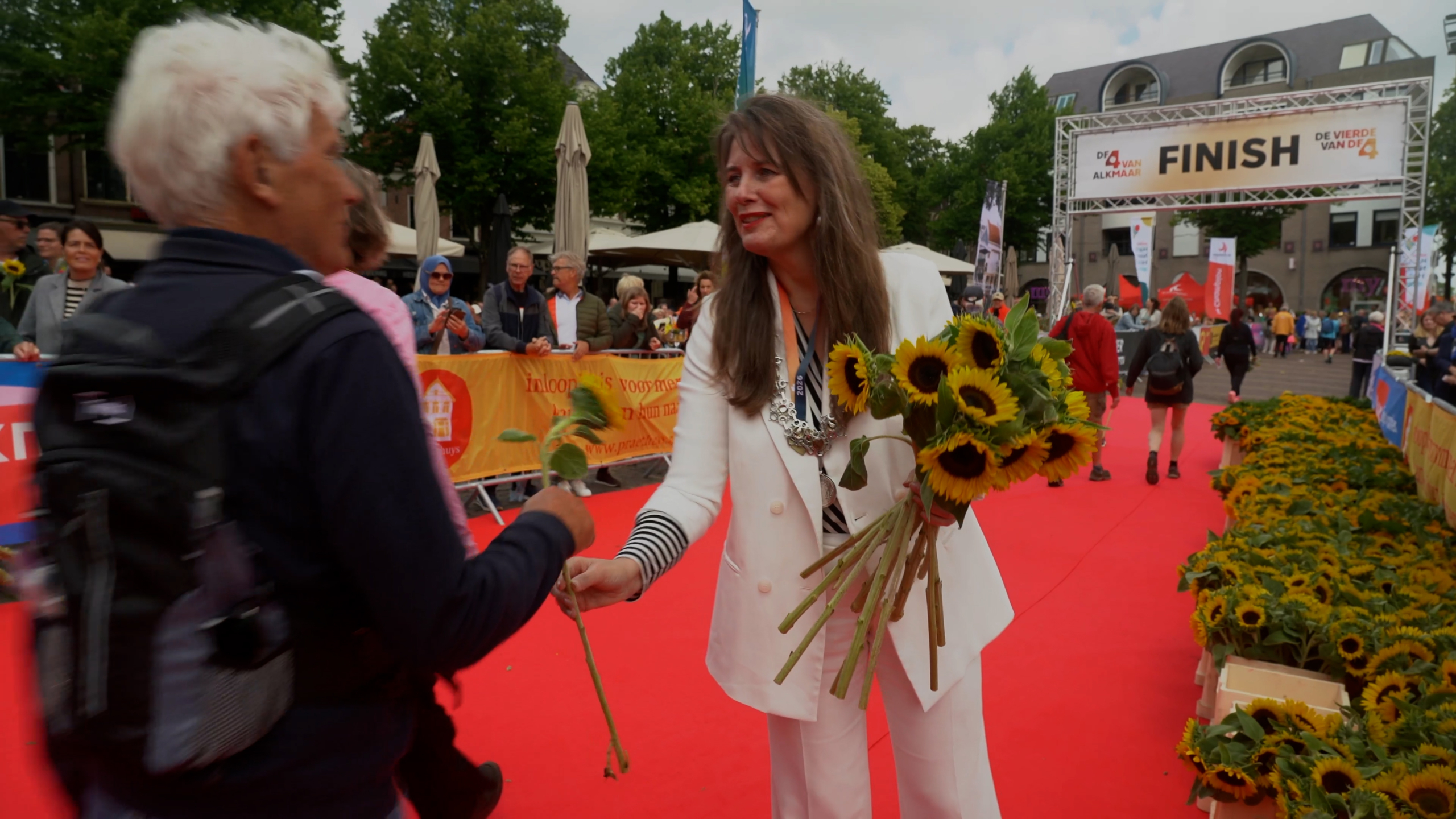 Een vrouw in een witte outfit geeft zonnebloemen aan een deelnemer bij de finish van een evenement met een rode loper.