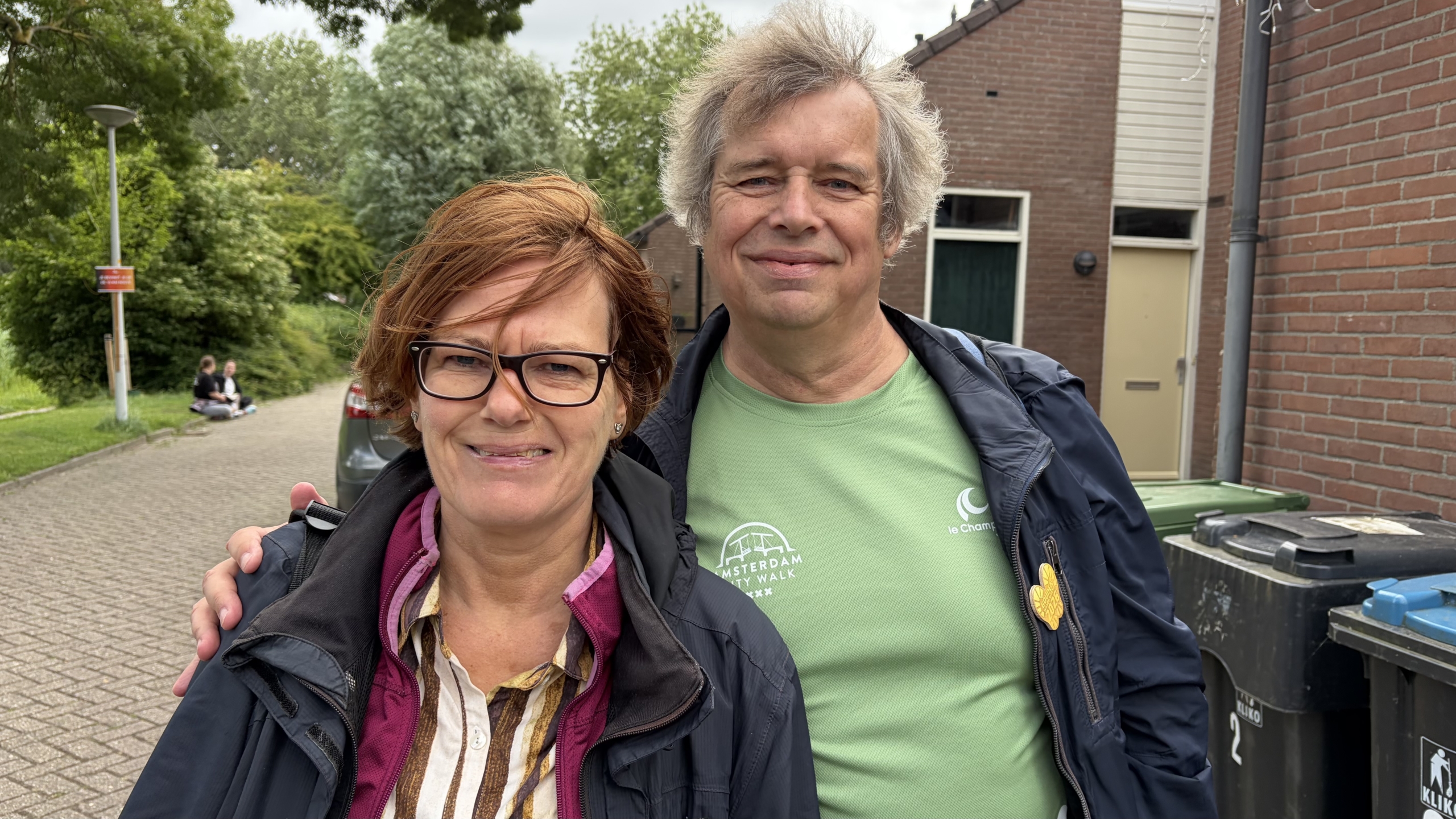 Een man en vrouw staan buiten dicht bij elkaar, beide glimlachend. Op de achtergrond is een woonwijk zichtbaar.