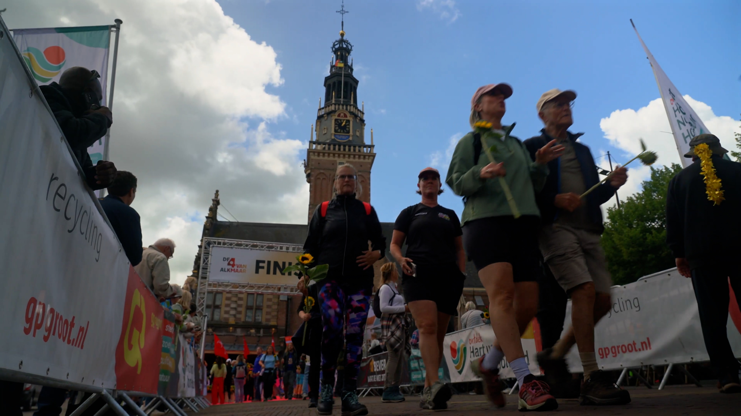 Mensen wandelen bij een finishlijn onder een blauwe lucht met wolken, met een toren op de achtergrond.