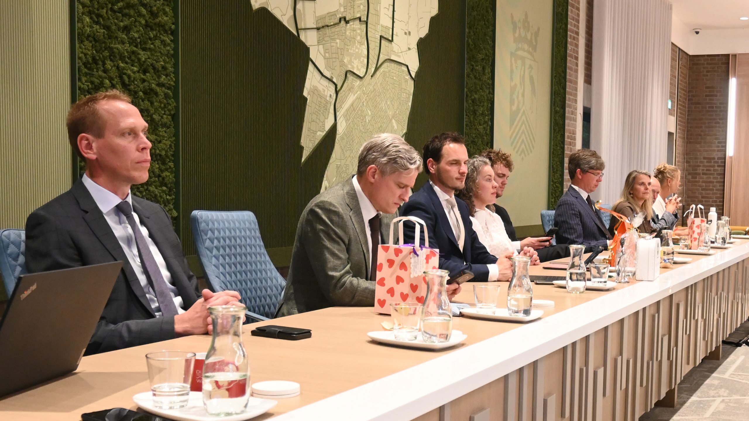 Mensen zitten aan een tafel in een vergaderruimte, omgeven door glazen en karaffen water, met een kaart van een regio op de muur.