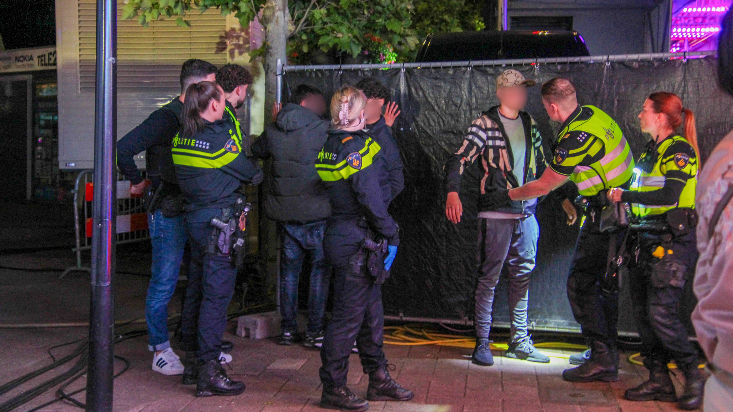 Politieagenten controleren twee mannen tegen een hek in een stedelijke omgeving.