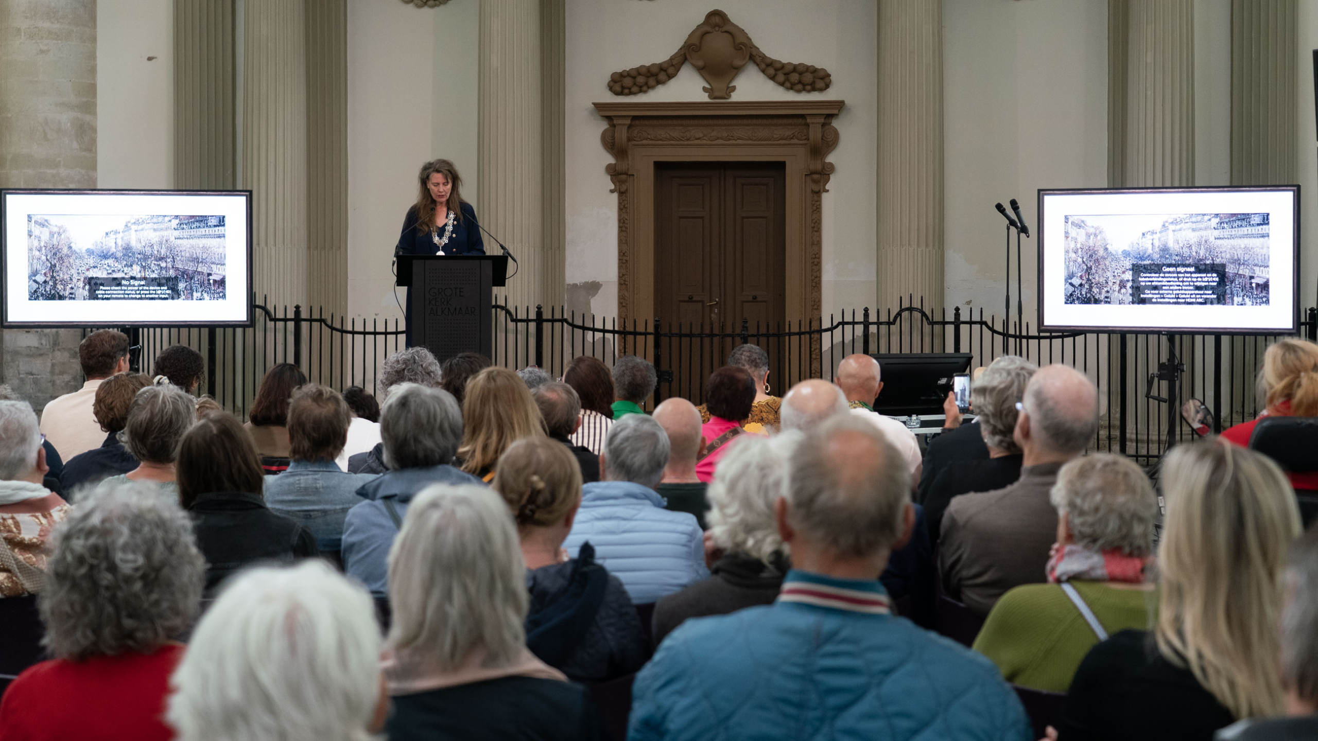 Een spreker presenteert voor een publiek in de Grote Kerk Alkmaar, met dia's op twee schermen.