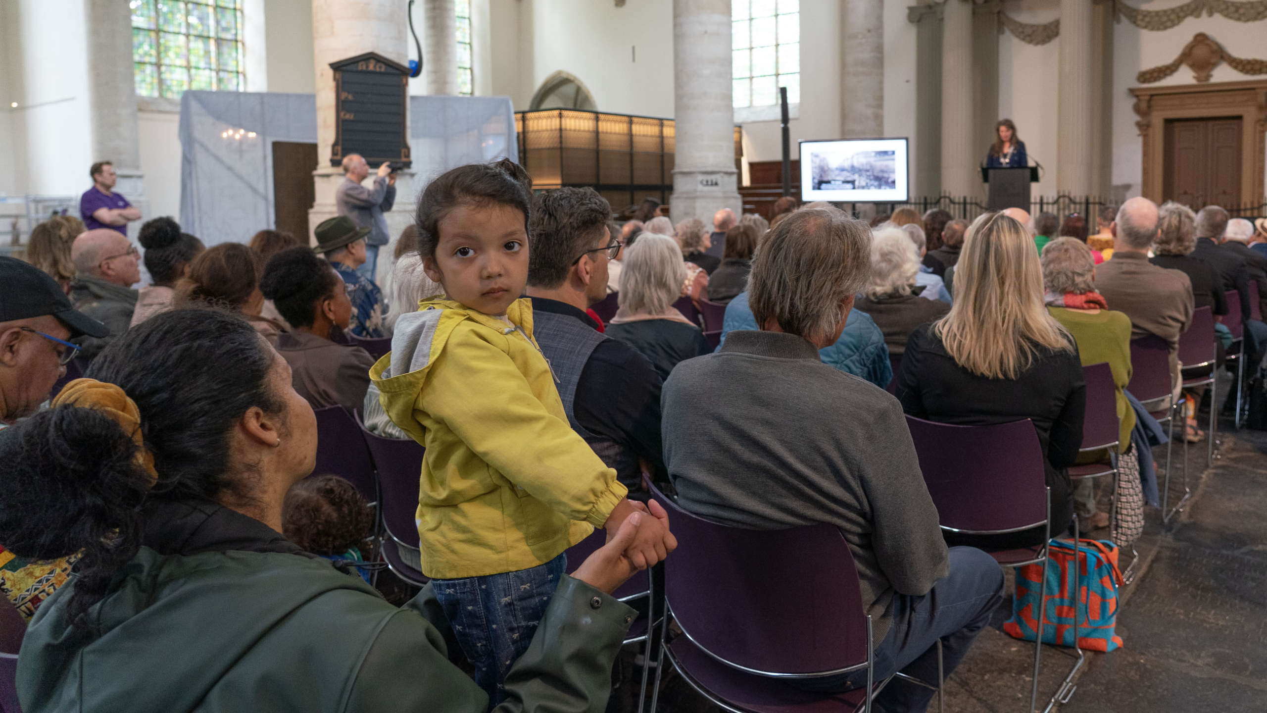 Een jong meisje in gele jas kijkt naar de camera terwijl ze de hand vasthoudt van een volwassene tijdens een presentatie in een kerk.