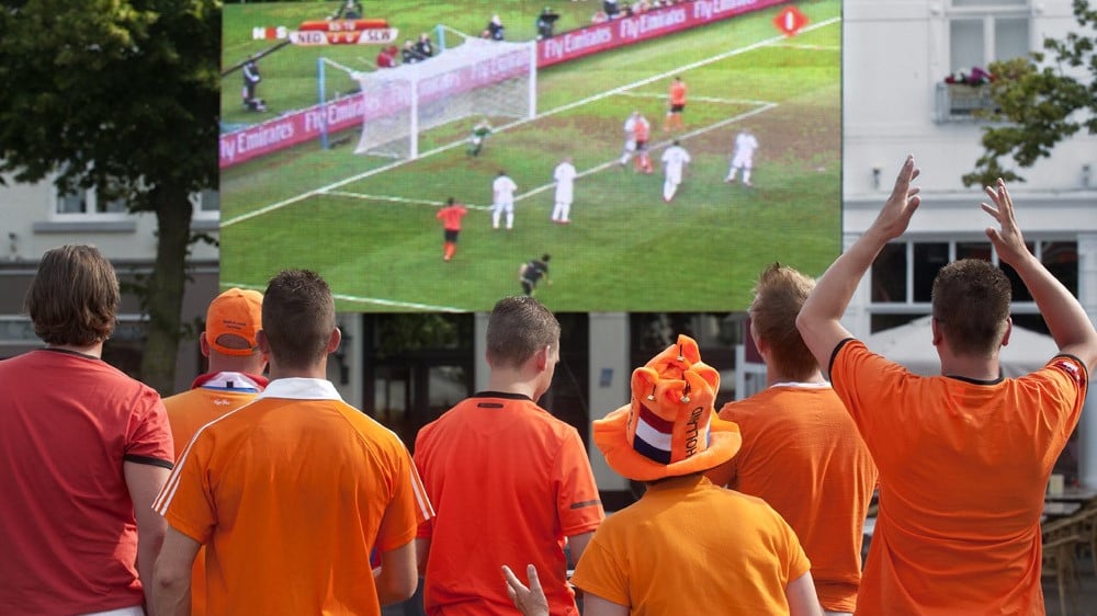 Voetbalfans in oranje kijken naar een grote scherm waarop een wedstrijd wordt uitgezonden, midden in een juichmoment.