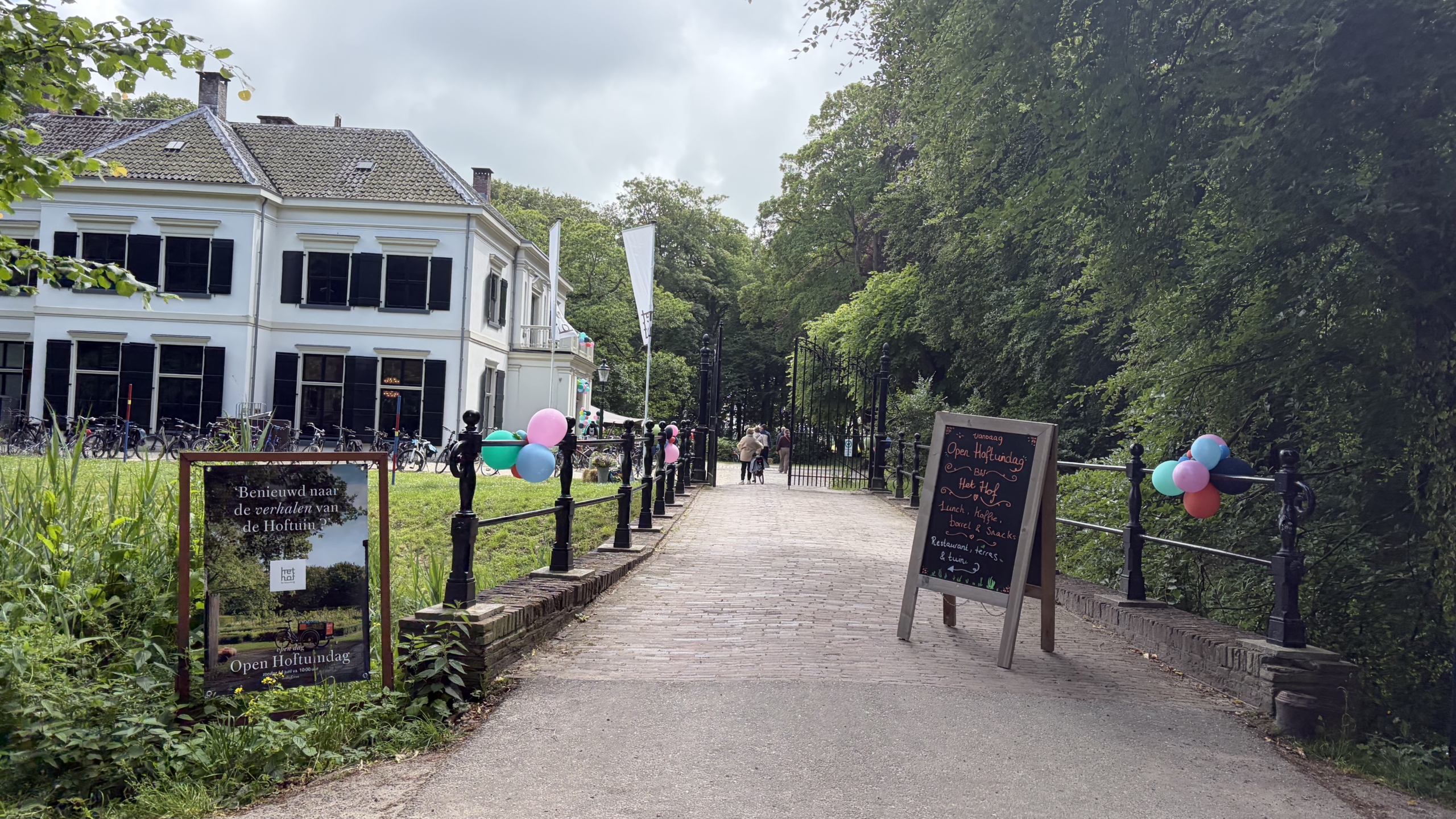 Een groot wit gebouw met zwarte luiken en fietsen ervoor, omringd door bomen, ballonnen en een bord met tekst over een "Open Hoftuindag."