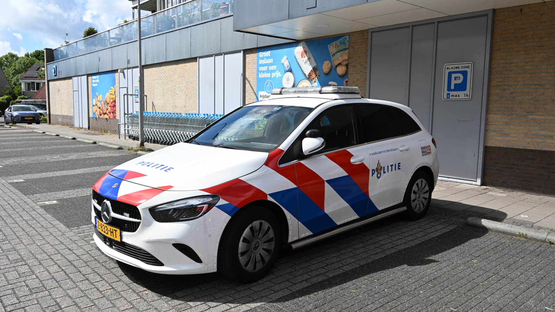 Een Nederlandse politieauto geparkeerd voor een supermarkt, met rood-blauwe strepen en "Politie" op de zijkant.