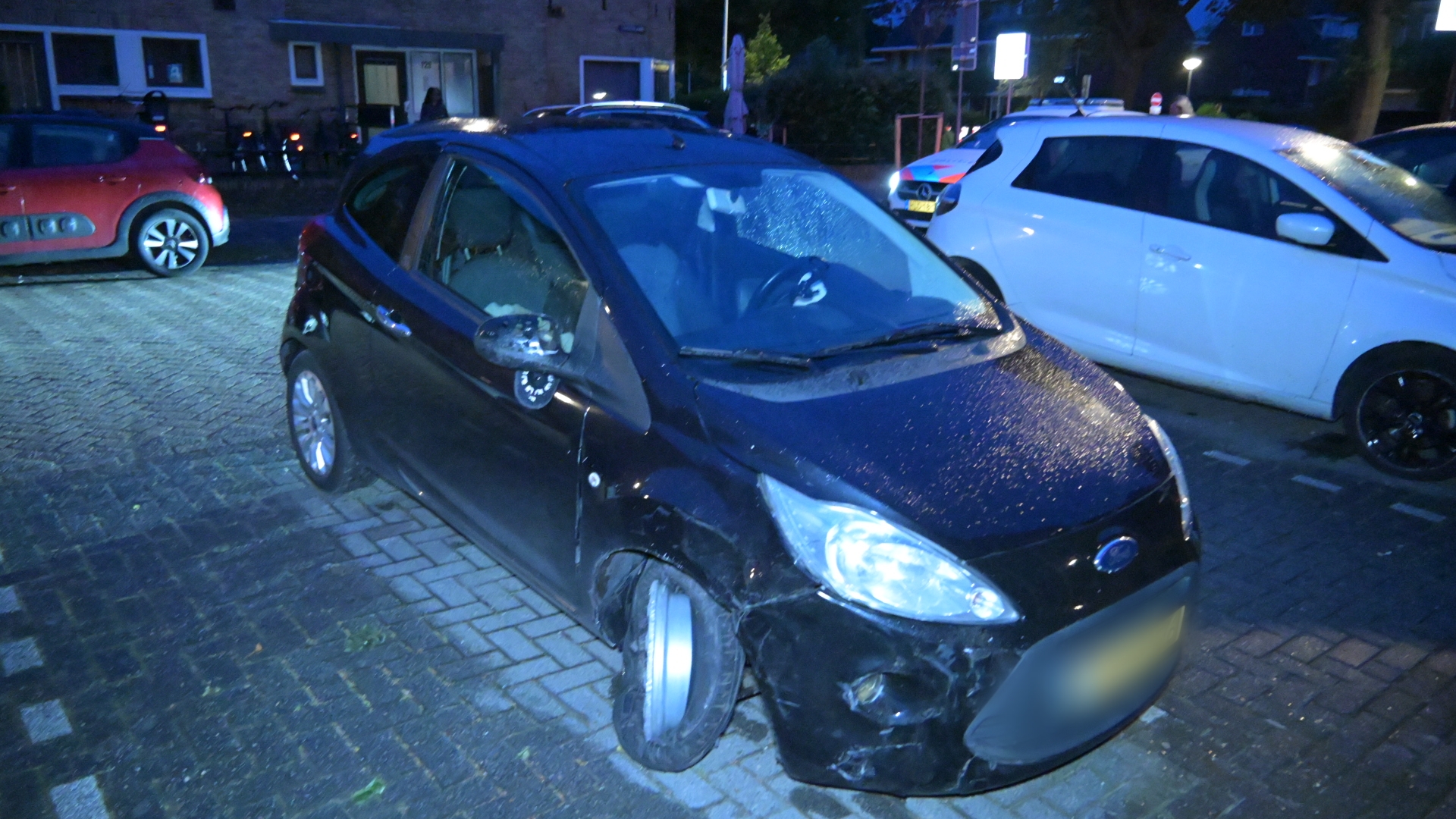 Een zwarte auto zonder linkervoorwiel staat beschadigd geparkeerd op een natte straat in het donker.