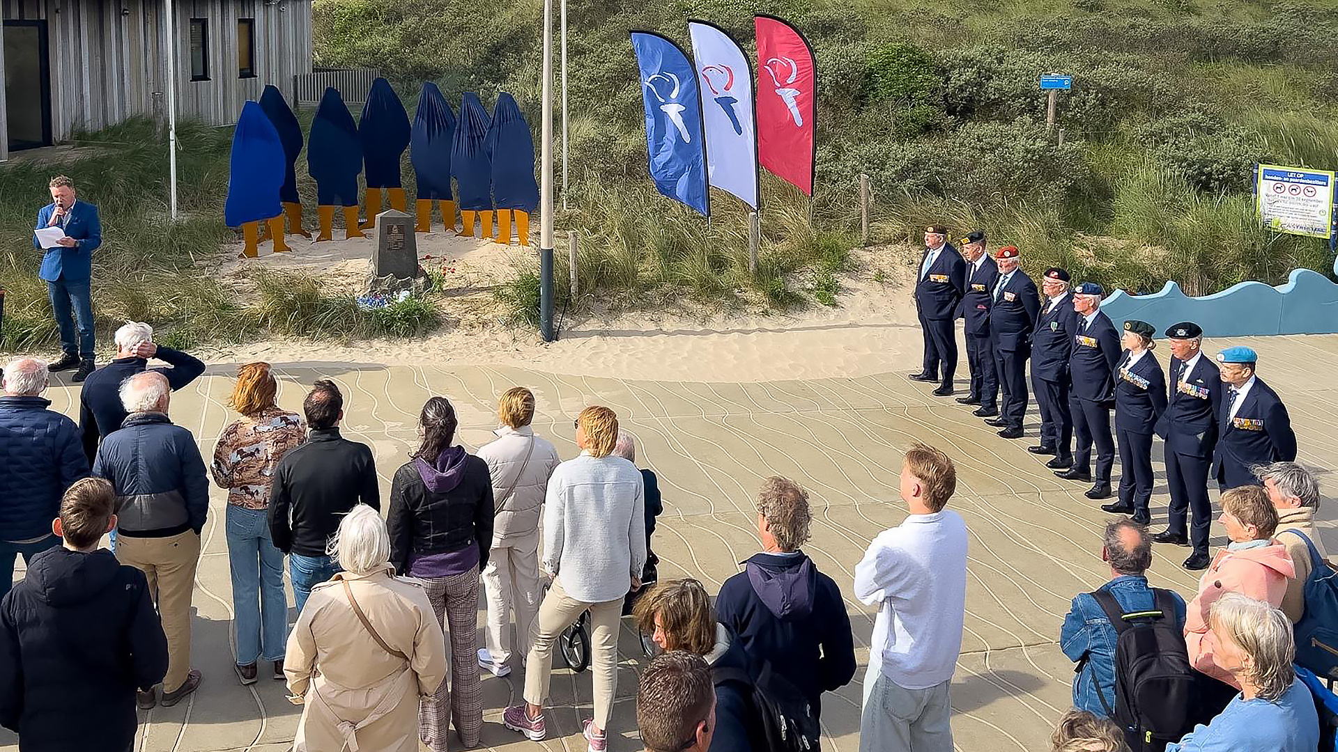 Mensen nemen deel aan een ceremonie buiten bij een monument, met verschillende vlaggen en uniformen.