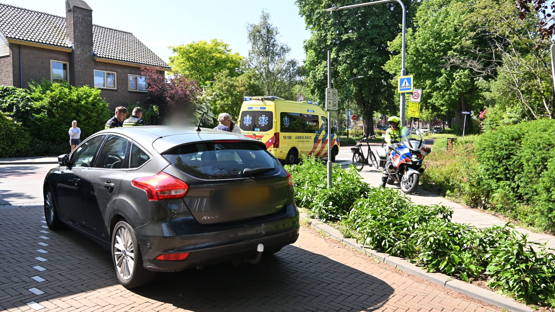 Politie en ambulance bij een incident met een zwarte auto op een zonnige dag, met een motoragent op de stoep.
