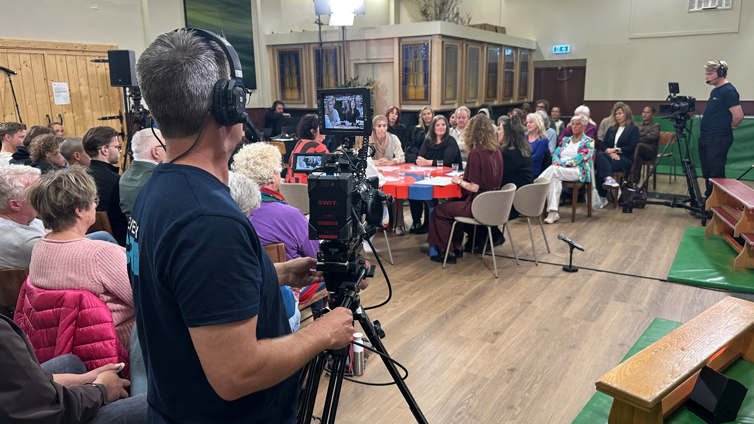 Een opname van een gesprek in een zaal, met mensen rond een tafel en een cameraman op de voorgrond.