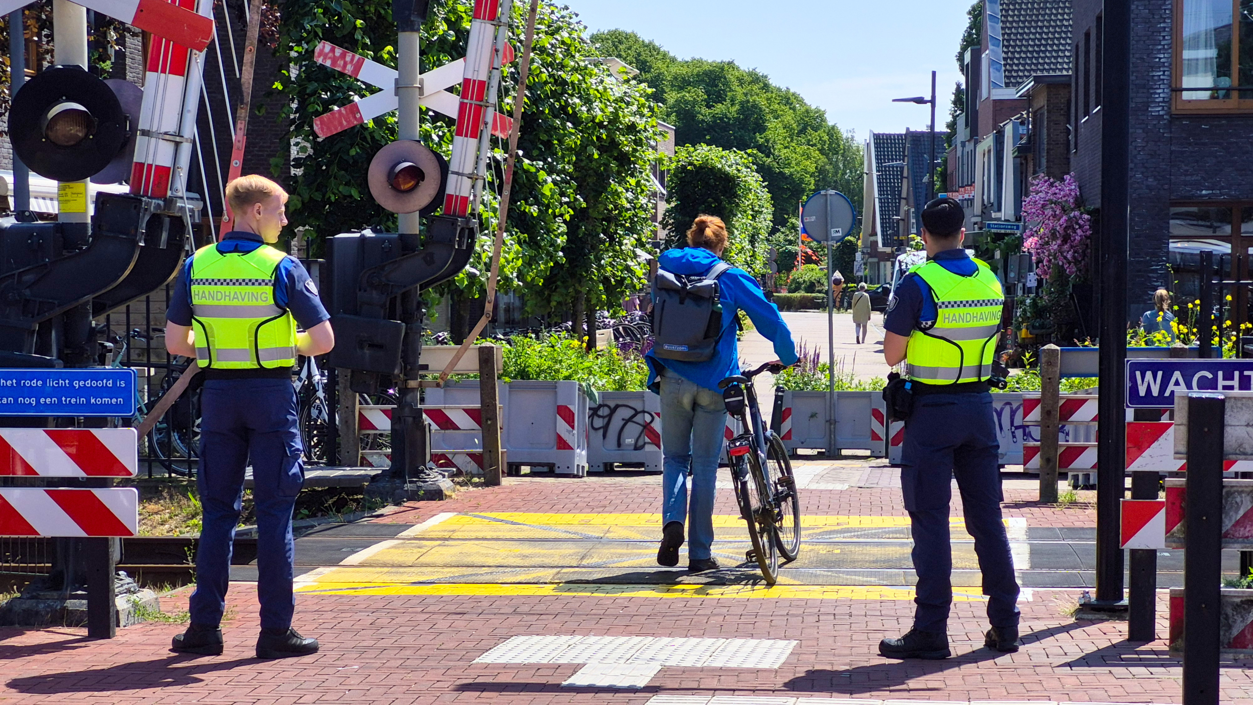 Twee handhavers bewaken een spoorwegovergang terwijl een fietser met fiets passeert.