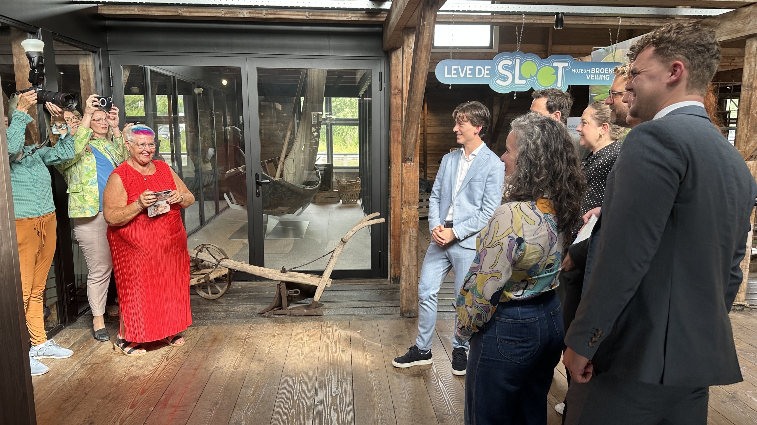 Groep mensen poseert lachend voor enkele fotografen in een houten gebouw met een bord met de tekst "Leve de Sloot".