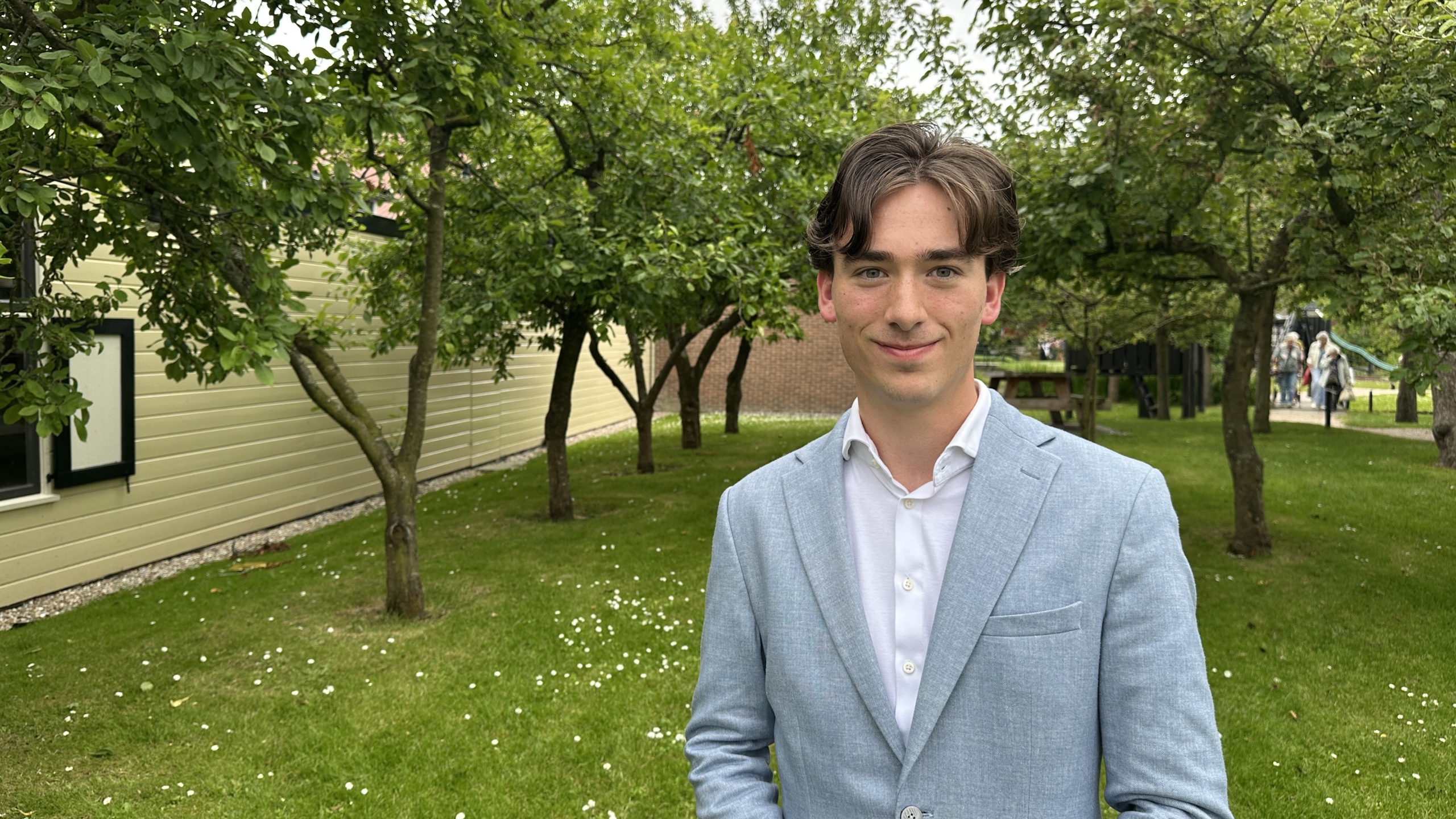 Een jonge man in een lichtblauw jasje staat glimlachend buiten in een boomrijke omgeving.