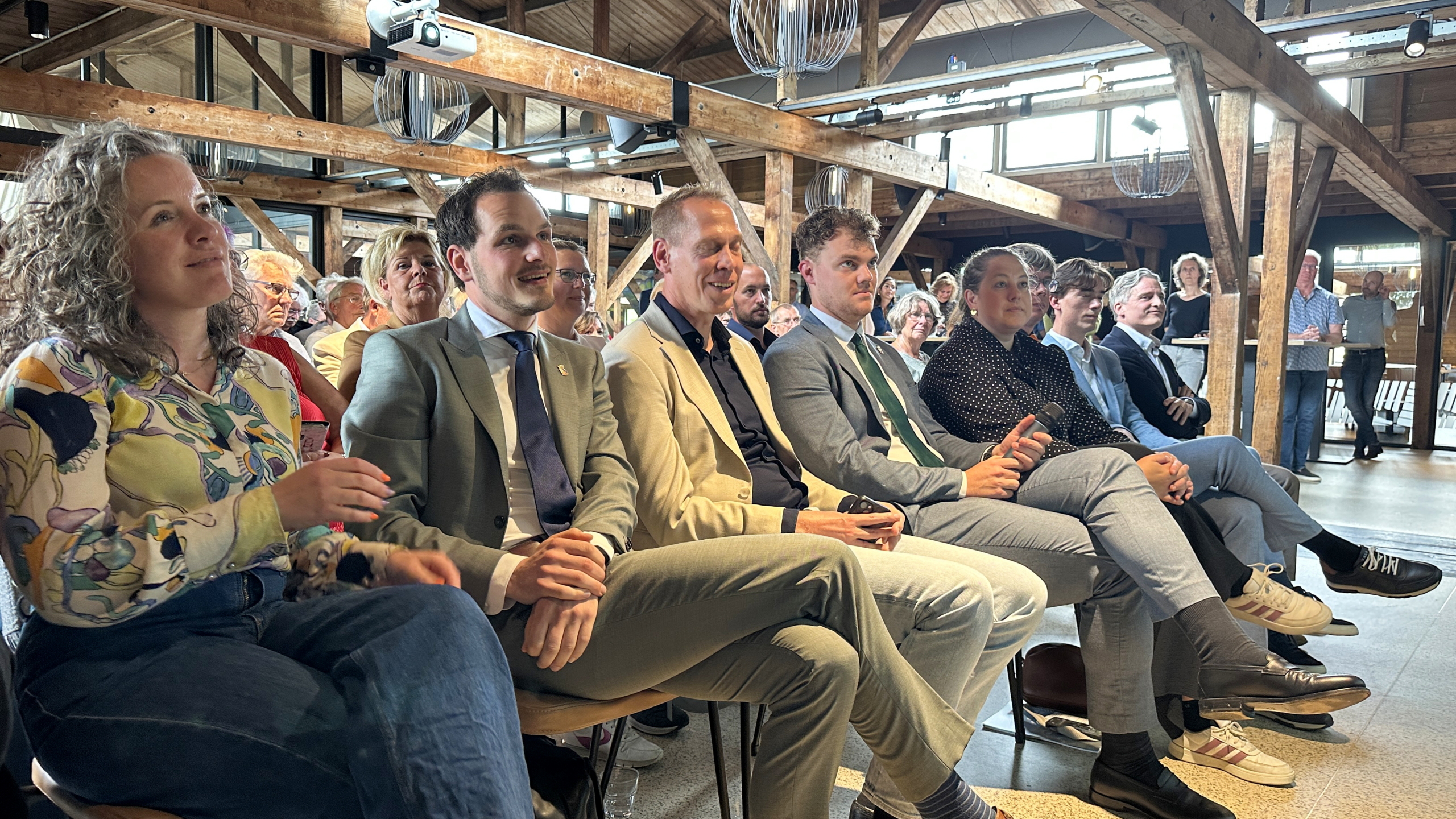 Een groep mensen zit aandachtig te luisteren in een zaal met een houten interieur, enkele houden een microfoon vast.