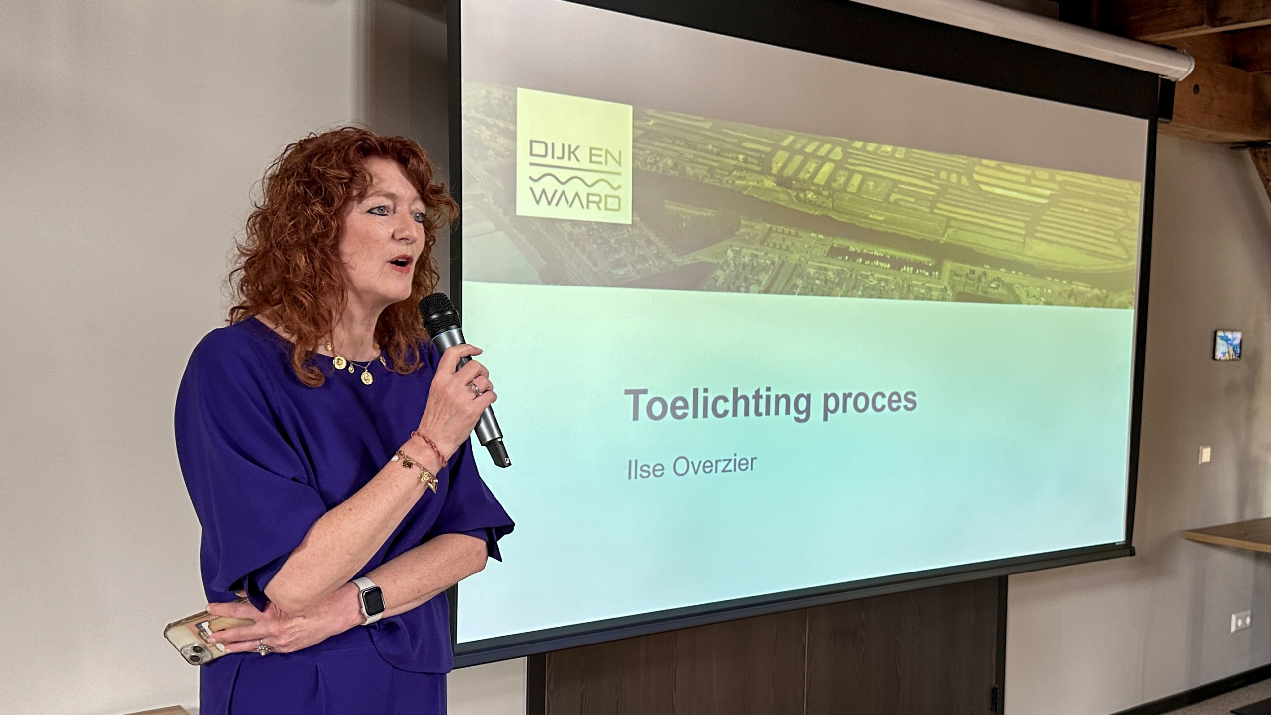 Vrouw met microfoon spreekt voor projector met presentatie "Toelichting proces" en logo "Dijk en Waard".